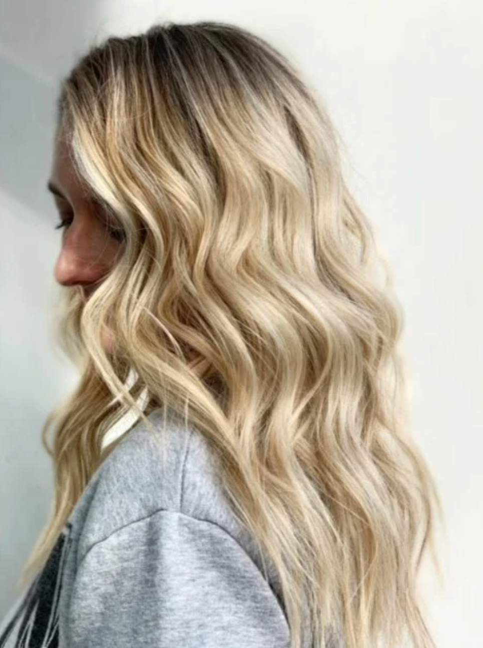 A woman with long, wavy blonde hair and beautiful blonde balayage highlights wearing a grey top, shown in profile against a plain background.