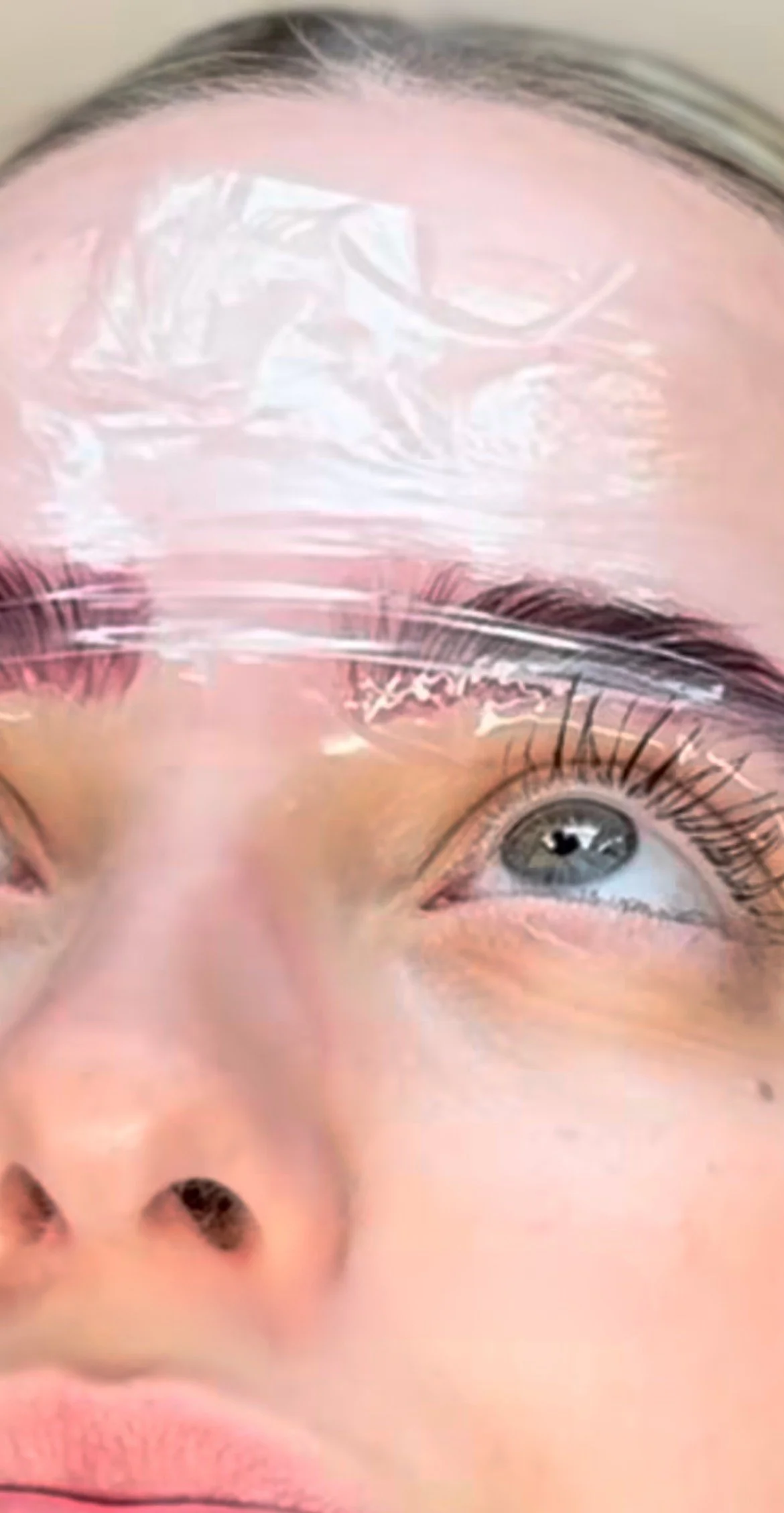 A woman receiving a brow lamination with a plastic sheet or mask over their face, partially covering their forehead, eyebrows, and eyes, with visible eyelashes and eye details.