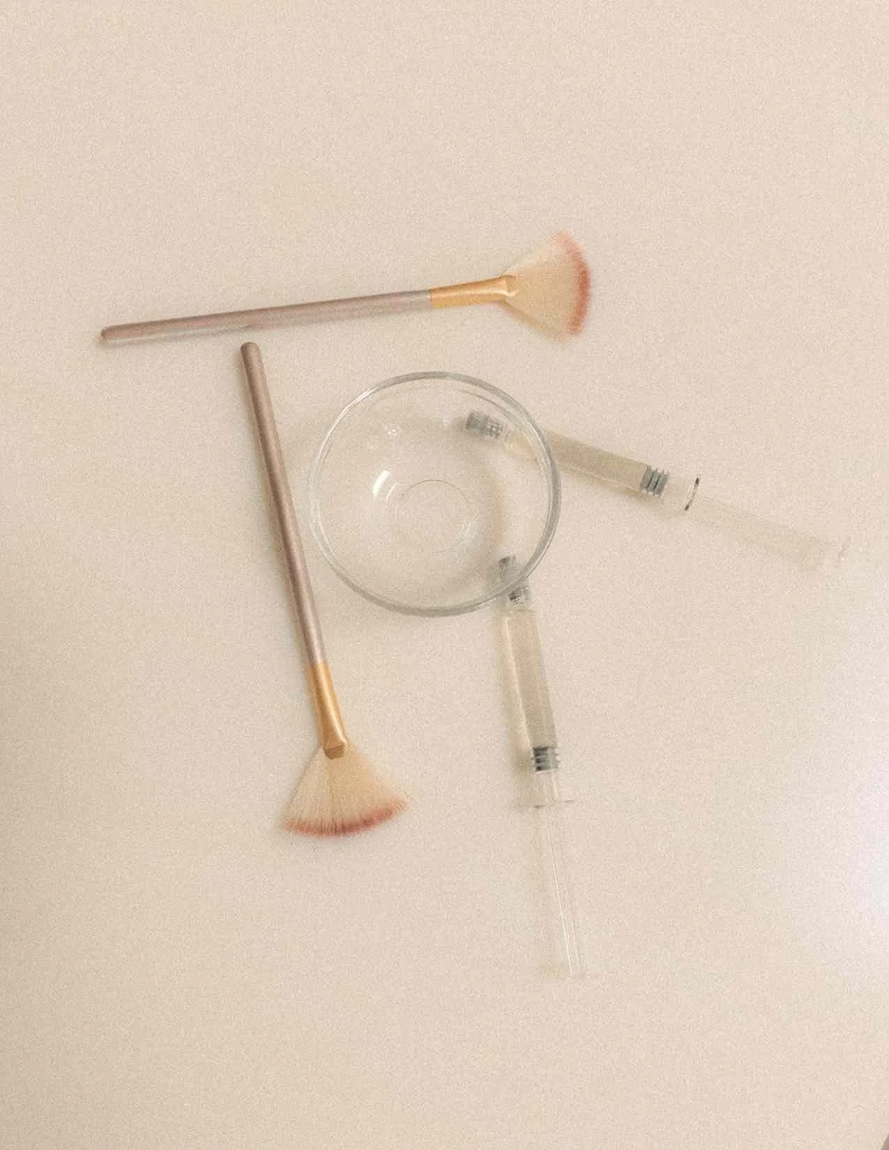 Cosmetic brushes, a small glass bowl, a syringe, and a test tube arranged on a beige surface.