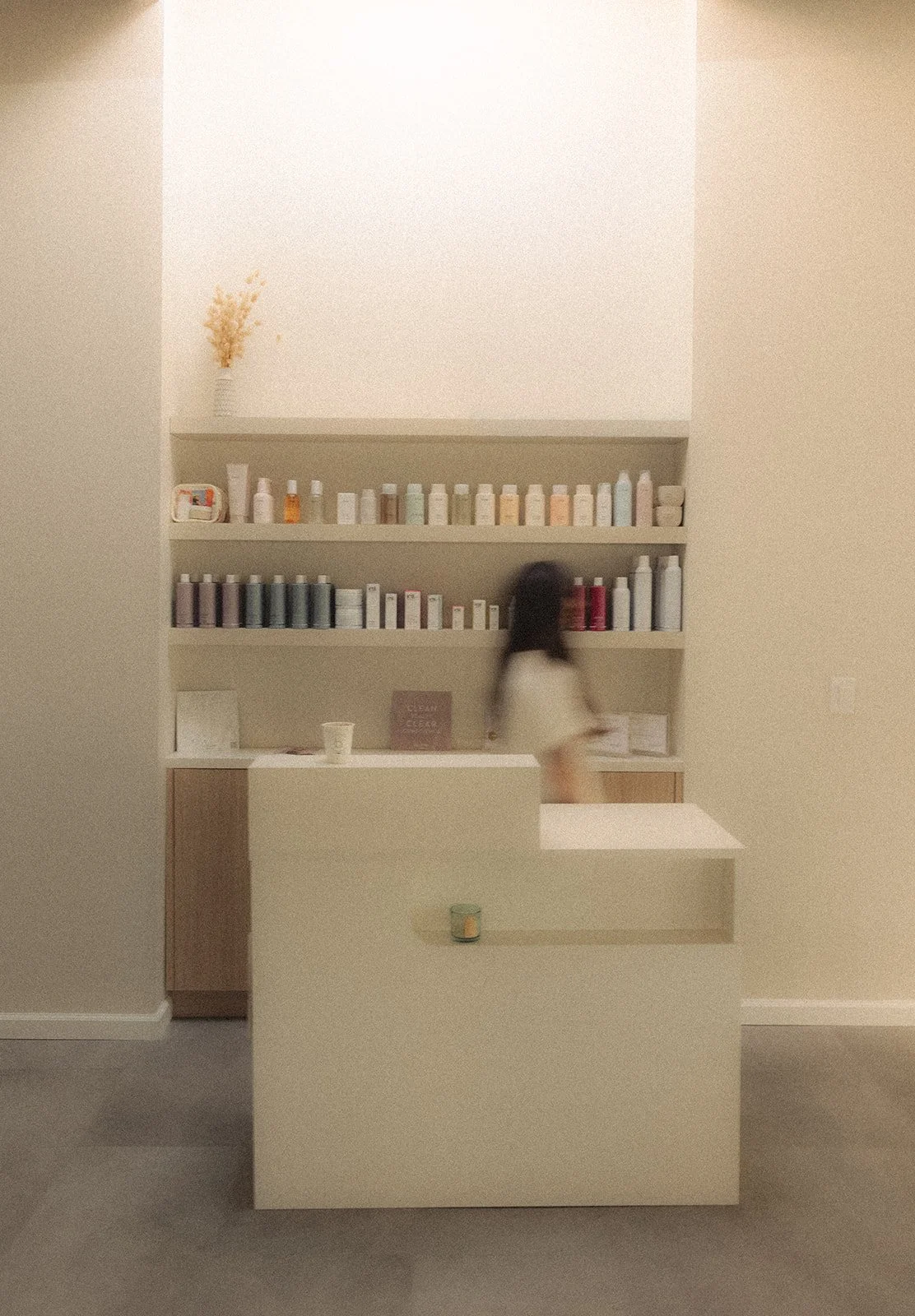 A woman walking past a beige reception desk with shelves of organized bottles behind it, in a minimalist interior space.