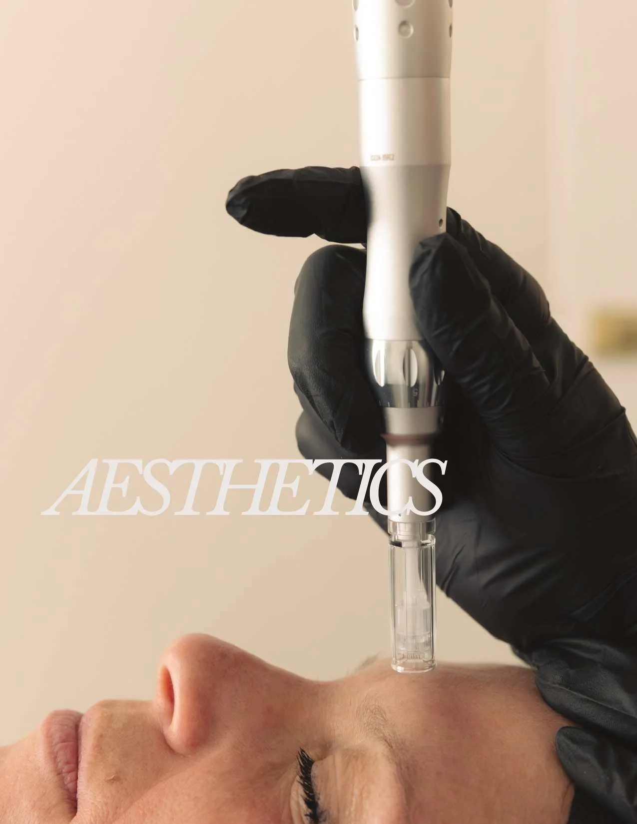 A person lying down with eyes closed receiving a cosmetic treatment from a medical professional wearing black gloves, holding a device near the person's forehead, with the word "AESTHETICS" overlayed on the image.