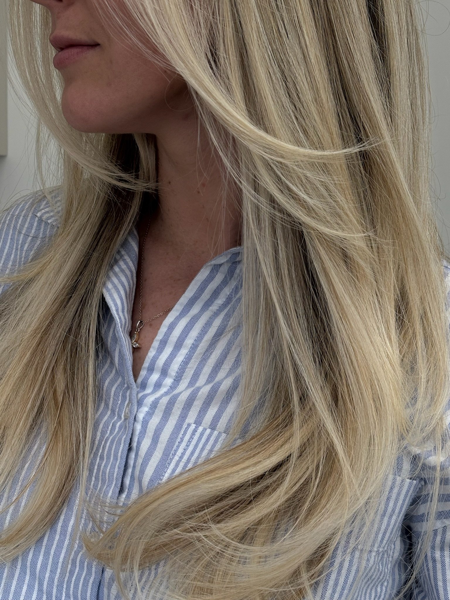 Close-up of a woman with long, wavy blonde hair, wearing a light blue, striped button-up shirt and a delicate necklace, with her face partially visible, turned slightly to the side.