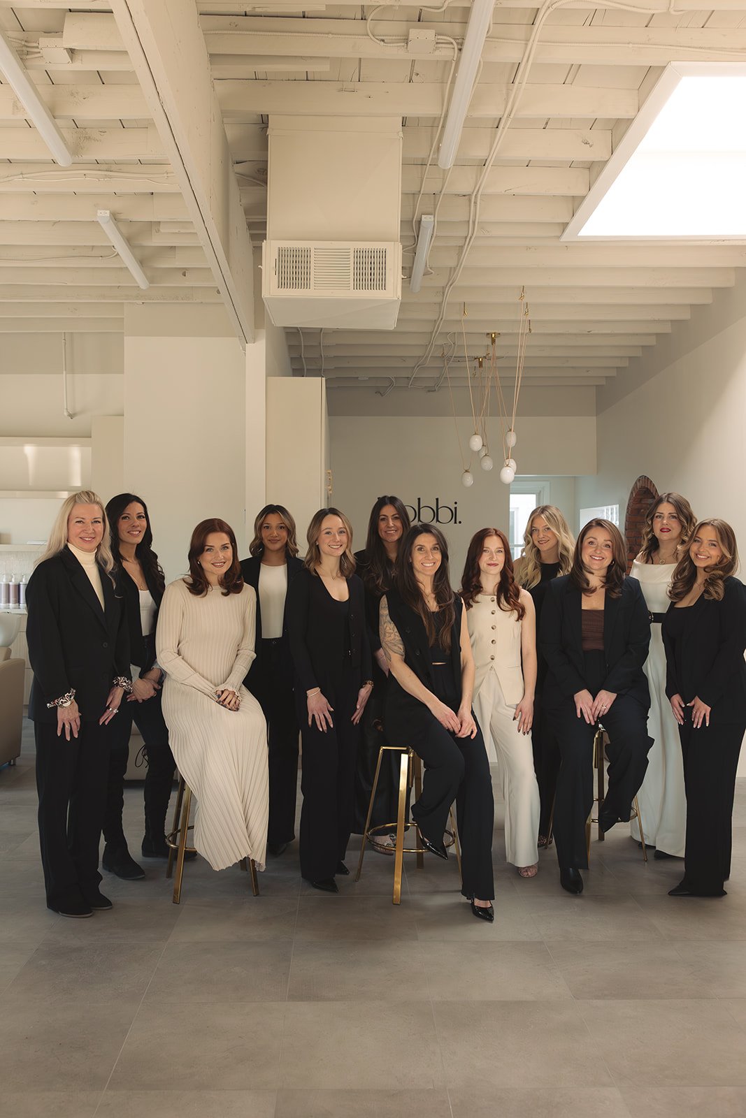 Bobbi staff, Grosse Pointe Premier Salon, dressed in professional attire standing and sitting together in a modern, well-lit interior space with a white ceiling and minimalist decor.