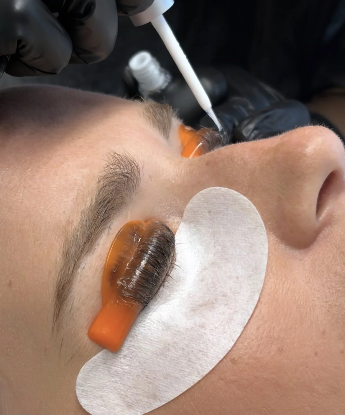 Close-up of a woman receiving Lash lift and tint, with protective eye patches and a technician applying adhesive with a brush, wearing black gloves.