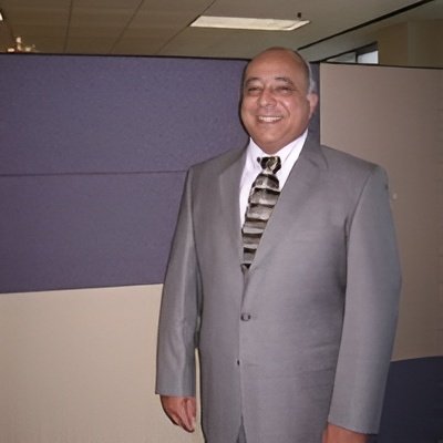 A smiling man in a gray suit and tie standing indoors in front of office cubicles.
