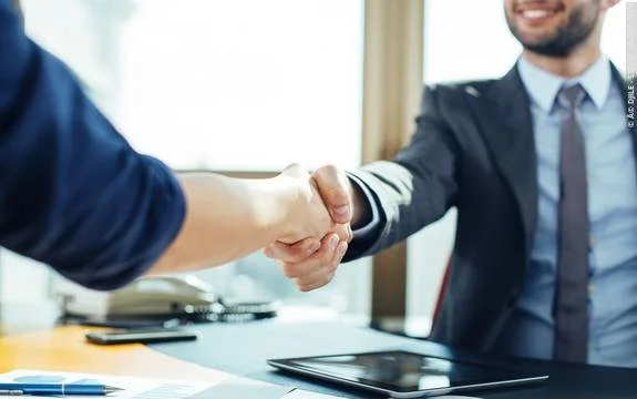 Two people shaking hands in a professional setting, with documents and a tablet on the table.