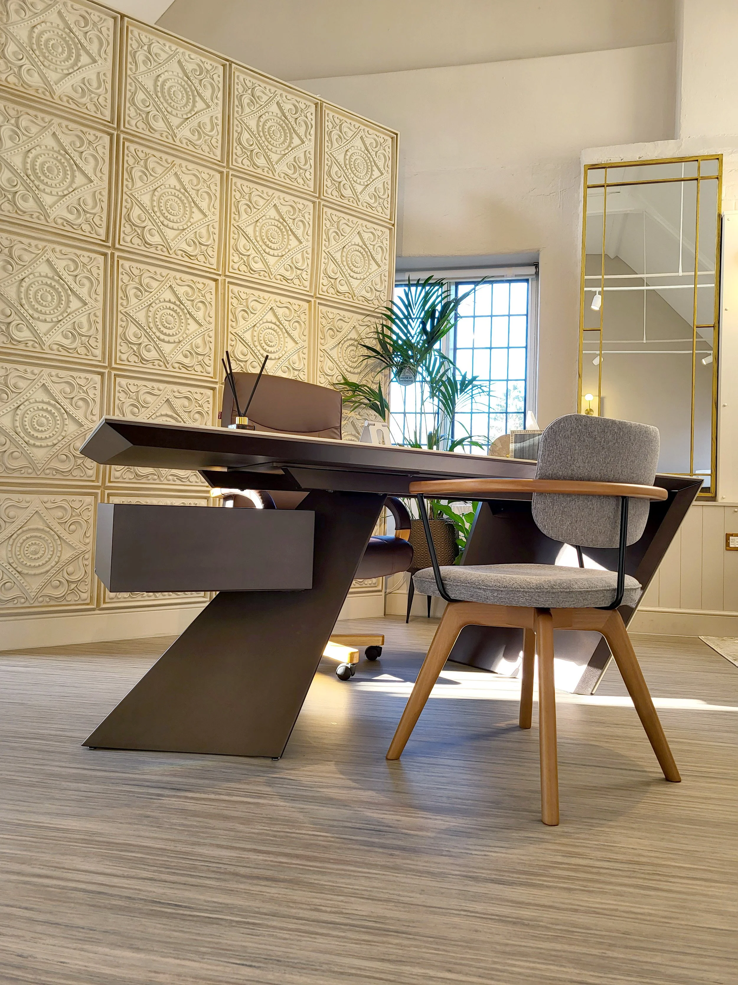 Modern office space with a desk, a grey upholstered chair, and a decorative wall with patterned panels. There are plants near a window, and a large mirror frame with a gold border on the wall.