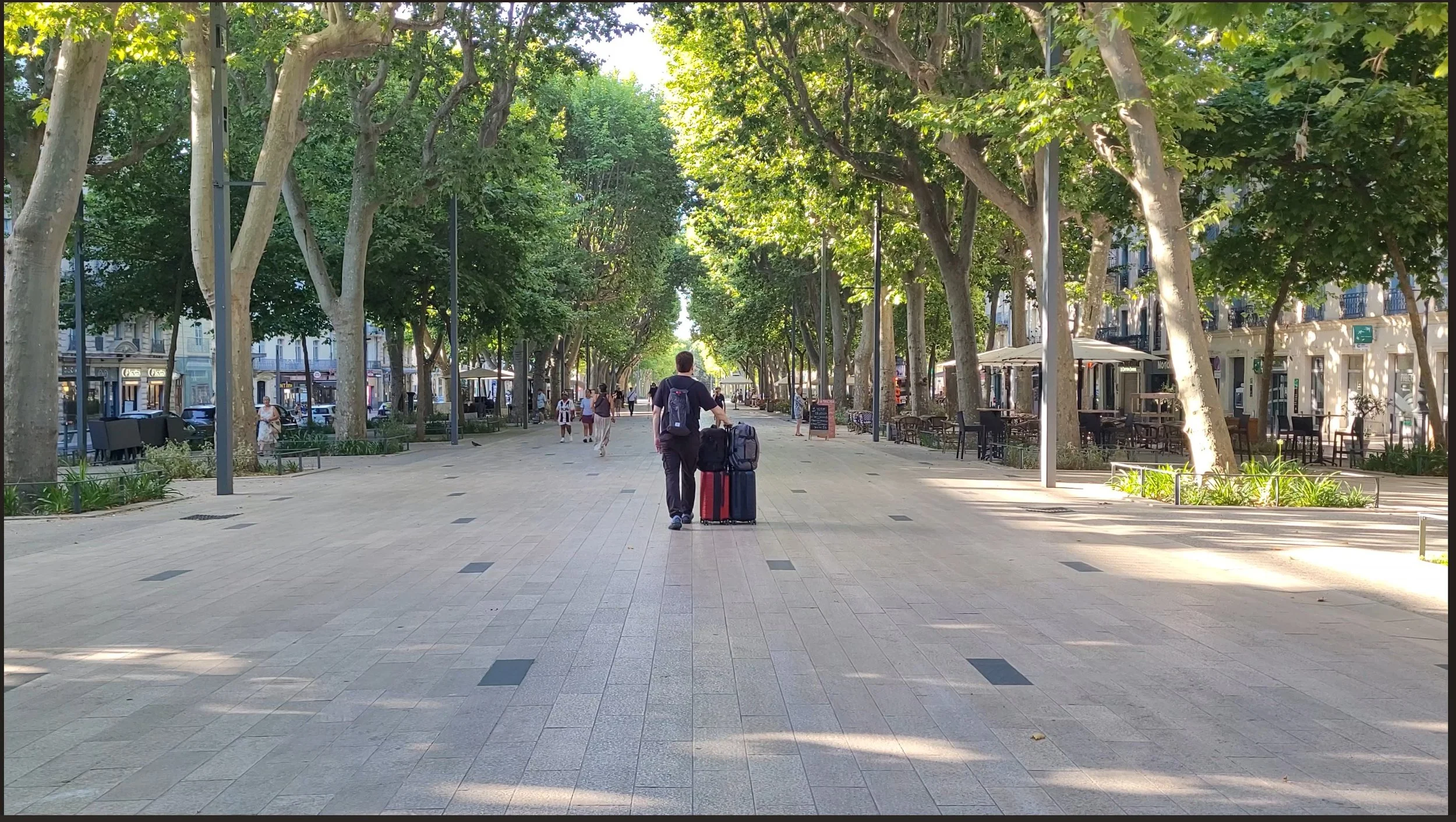 Allées Paul Riquet — broad promenade with plane trees, a traveler walking with luggage in dappled light