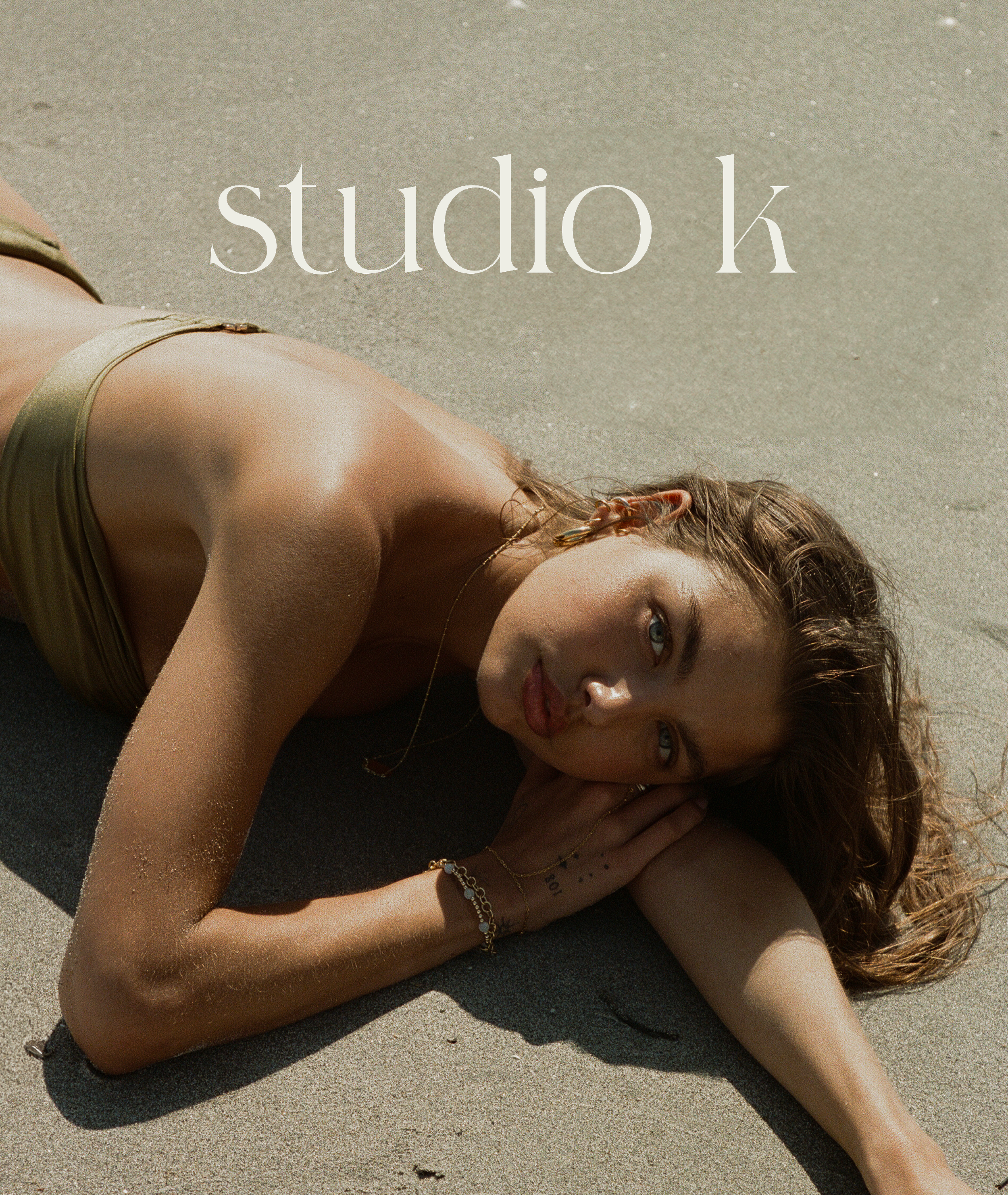 A woman with brown hair lying on sand at the beach, wearing a yellow bikini top and gold jewelry, gazing at the camera with her head resting on her arm.