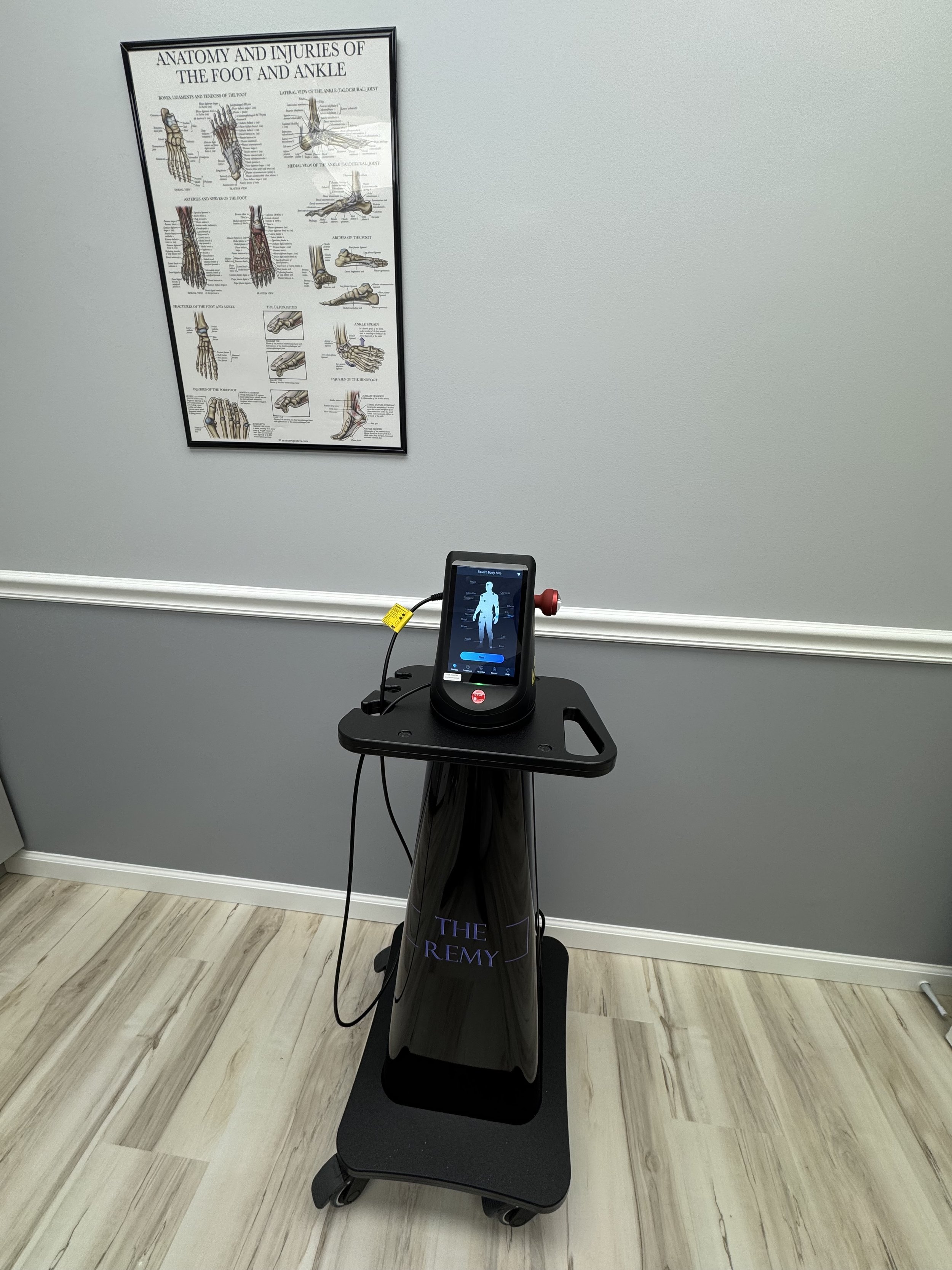 Remy Laser machine against a gray wall with an anatomical poster about foot and ankle injuries.