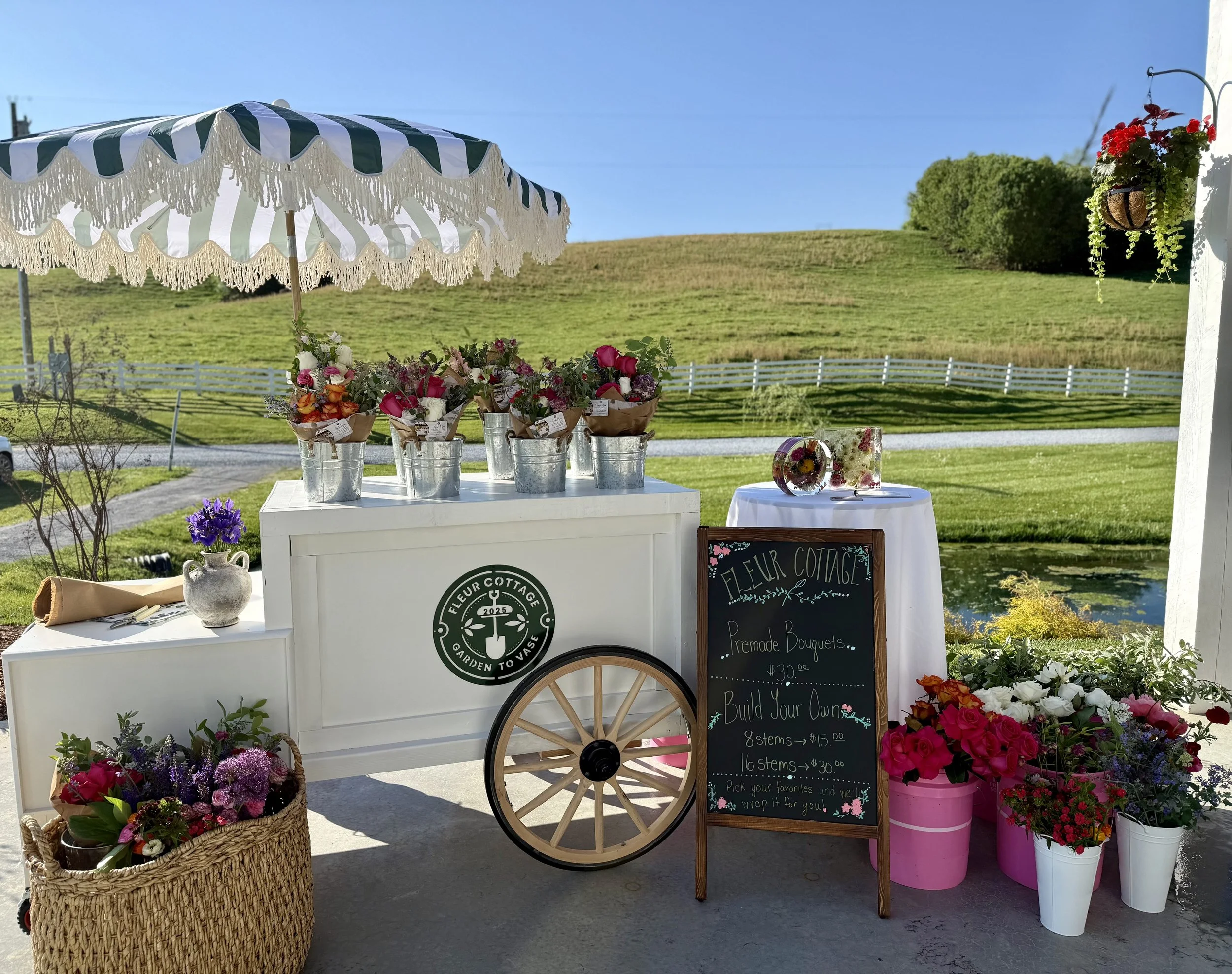 A flower cart titled 'Fleur Cottage' features colorful local fresh bouquets in buckets, a chalkboard sign with pricing, and decorative flowers in pots and baskets on a sunny day against a beautiful flower farm.