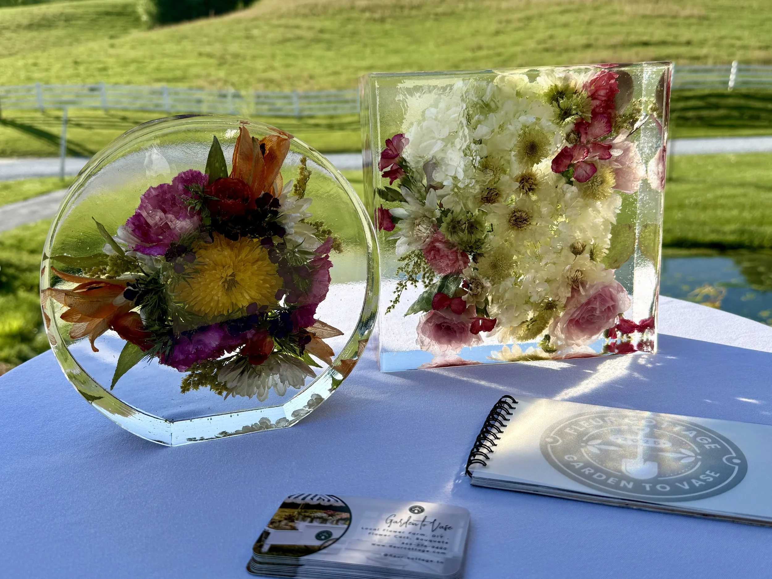 Floral preservation keepsakes in resin, one round and one rectangular, are placed on a table outdoors with a grassy hill and a white fence in the background. Preserved flowers from a special occasion.