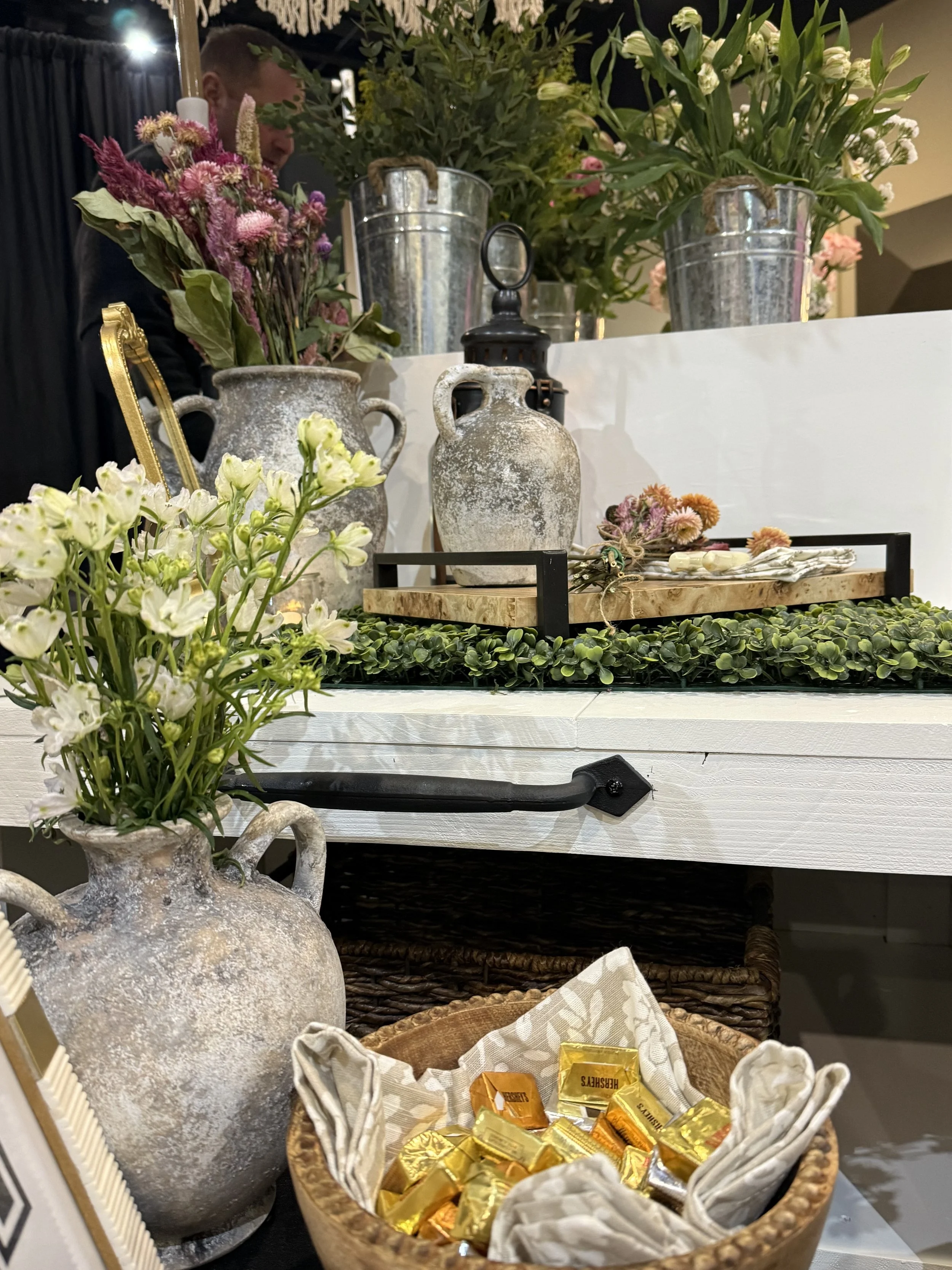 Decorative display with antique vases filled with pink, purple, and white flowers, green foliage, and a basket of wrapped candies. Example of a flower cart at a wedding, bridal shower, birthday, or other special event.