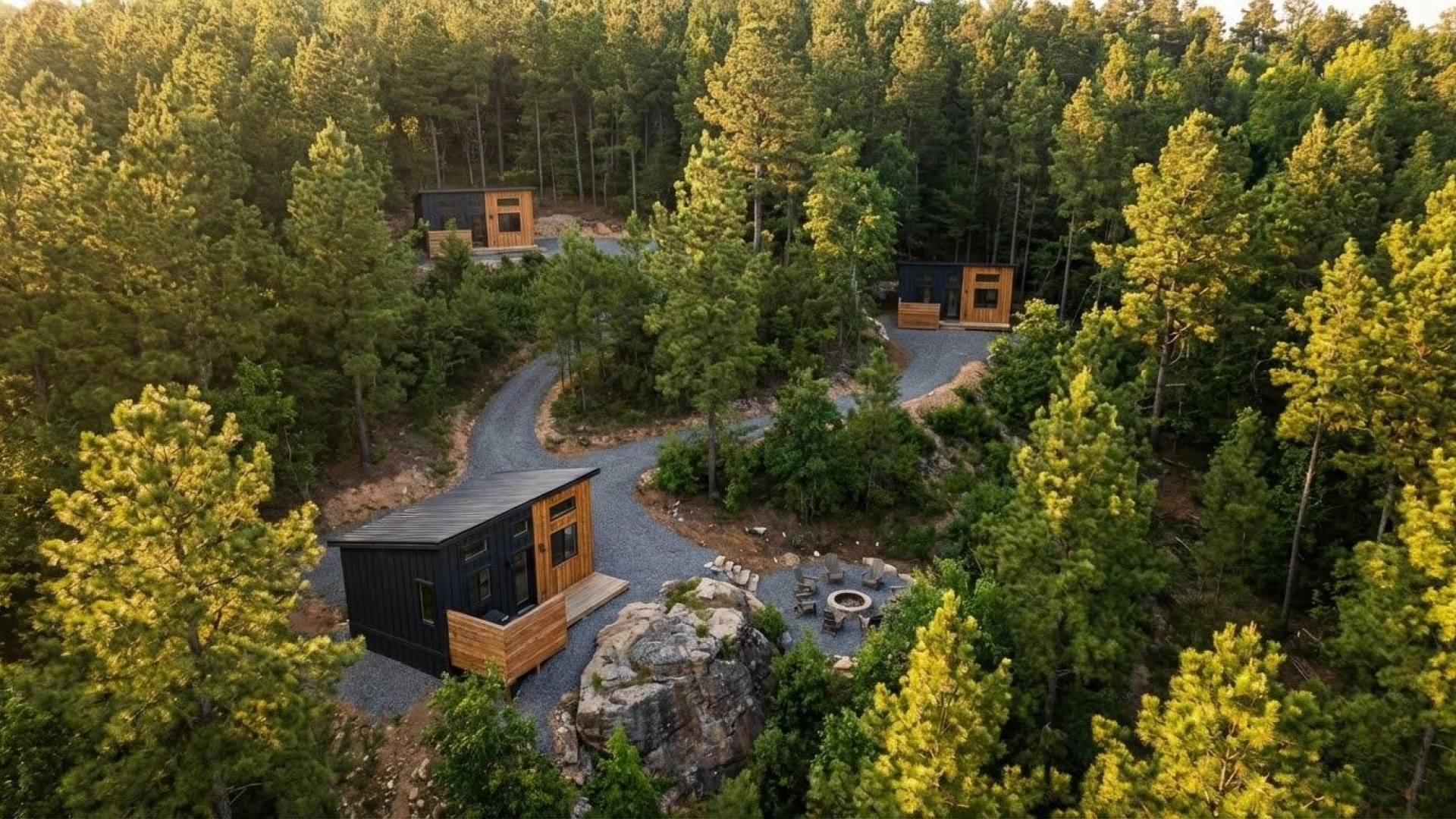 An aerial view of three small, modern cabins with contrasting black and natural wood siding, nestled in a dense green forest and connected by a winding gravel path, with a fire pit and seating area.