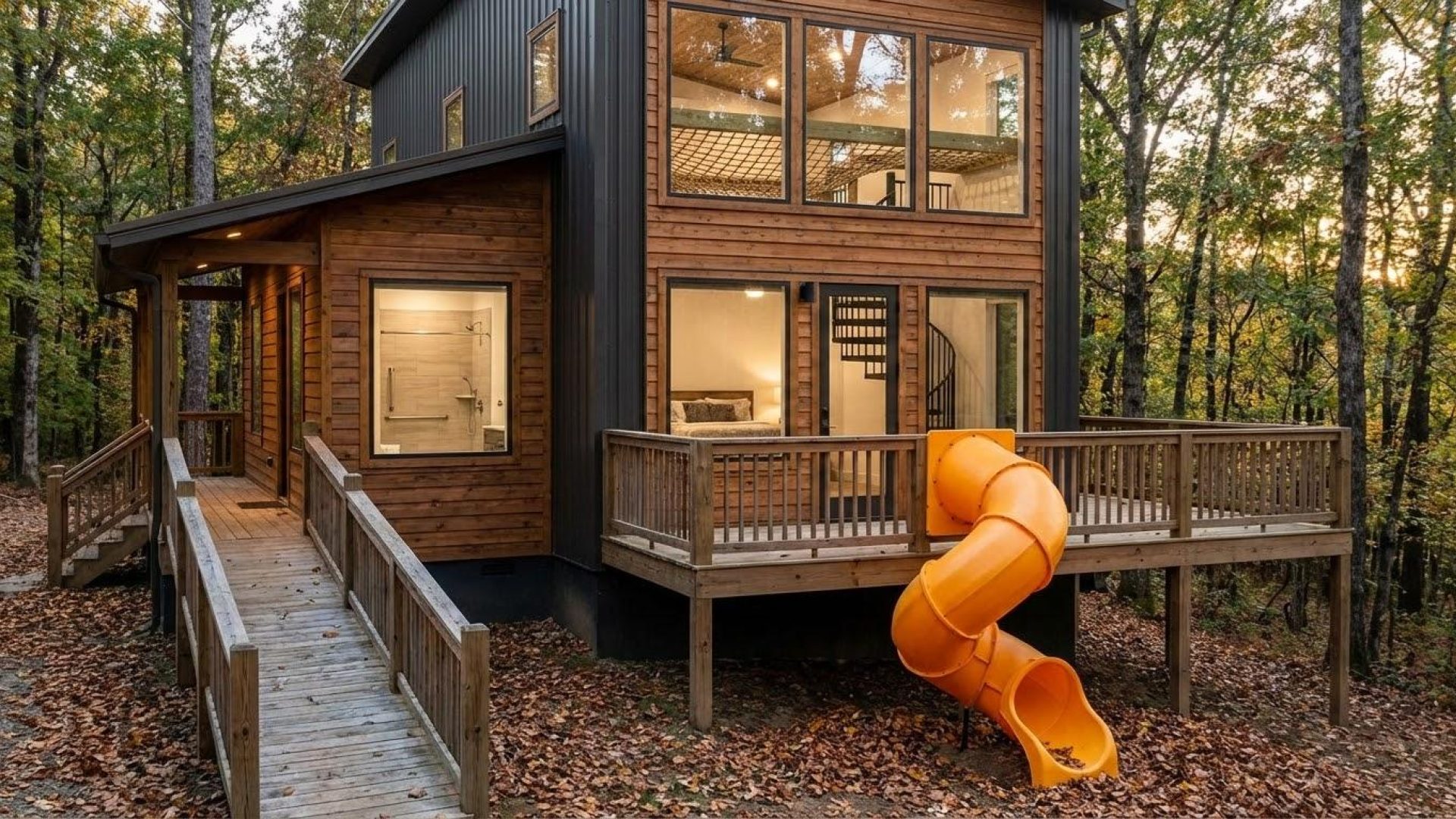 A modern two-story cabin with wood and dark metal siding features a prominent yellow spiral slide from the second-floor deck, an accessible wooden ramp leading to a side entrance, and large windows, all set within a dense forest.