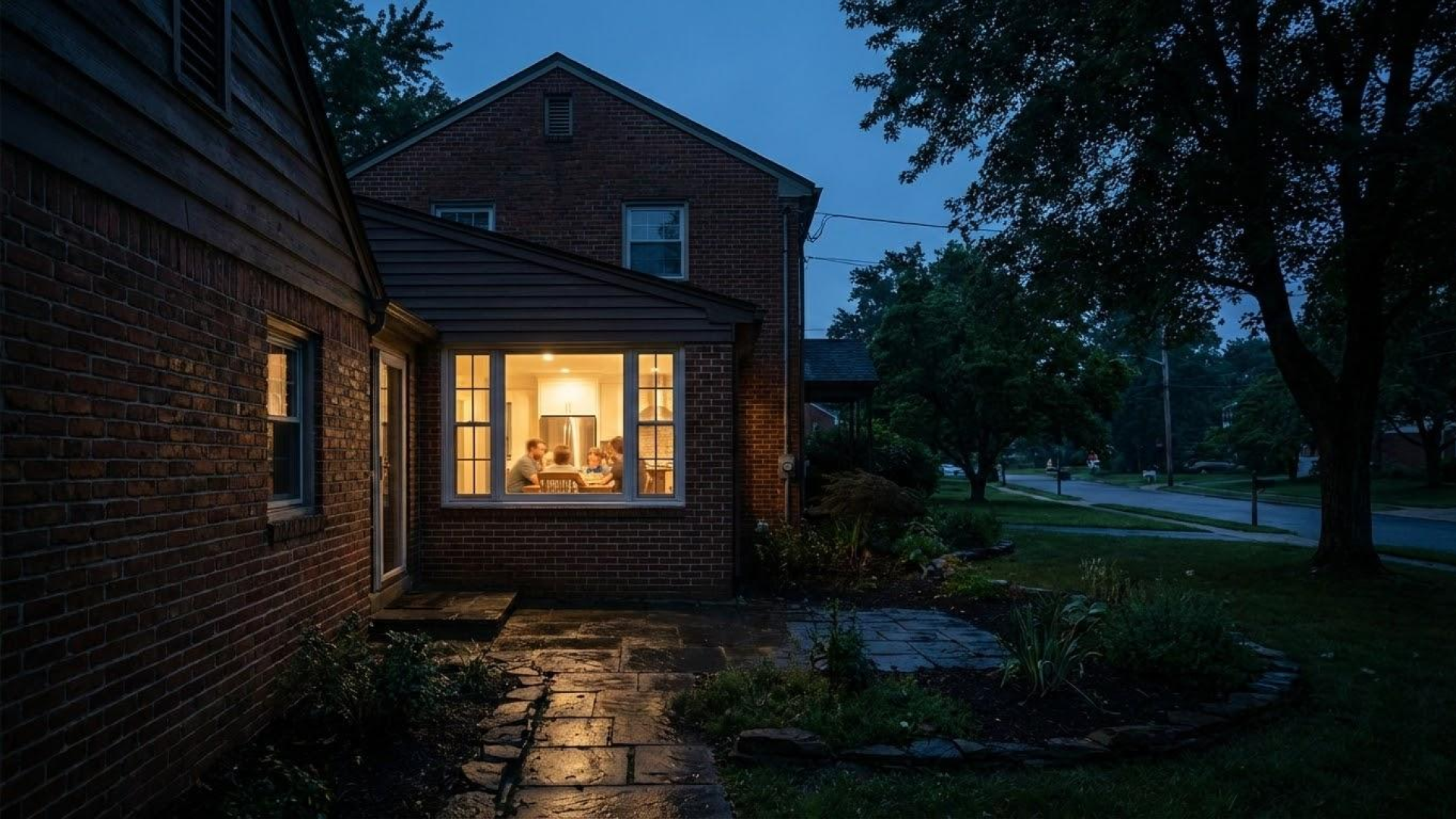 A view of a brick house at dusk with a brightly lit dining room visible through large windows, and a garden with trees and a driveway outside.