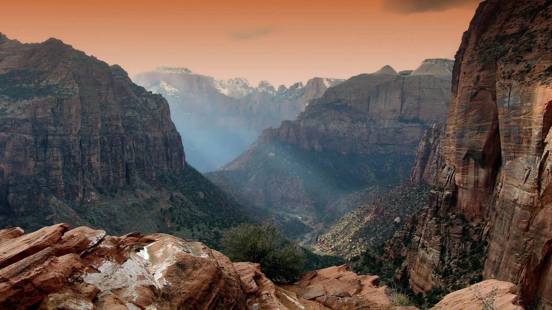 A scenic view of Zion Canyon with towering red and brown sandstone cliffs, some with snow, under a peach-colored sky at sunset or sunrise. All In For Utah celebrates the state's scenery and unique destinations.