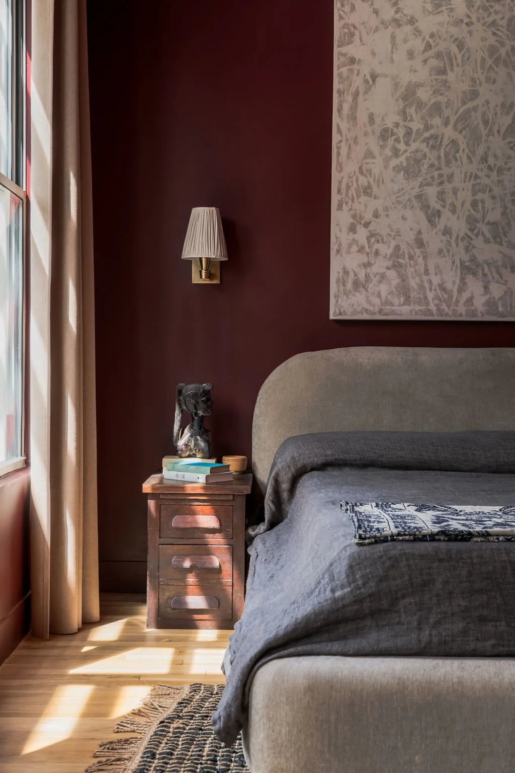 A moody bedroom with deep burgundy walls, a curved grey upholstered headboard, and charcoal linen bedding. A vintage wood nightstand holds stacked books and a dark sculptural bust beneath a brass pleated wall sconce.
