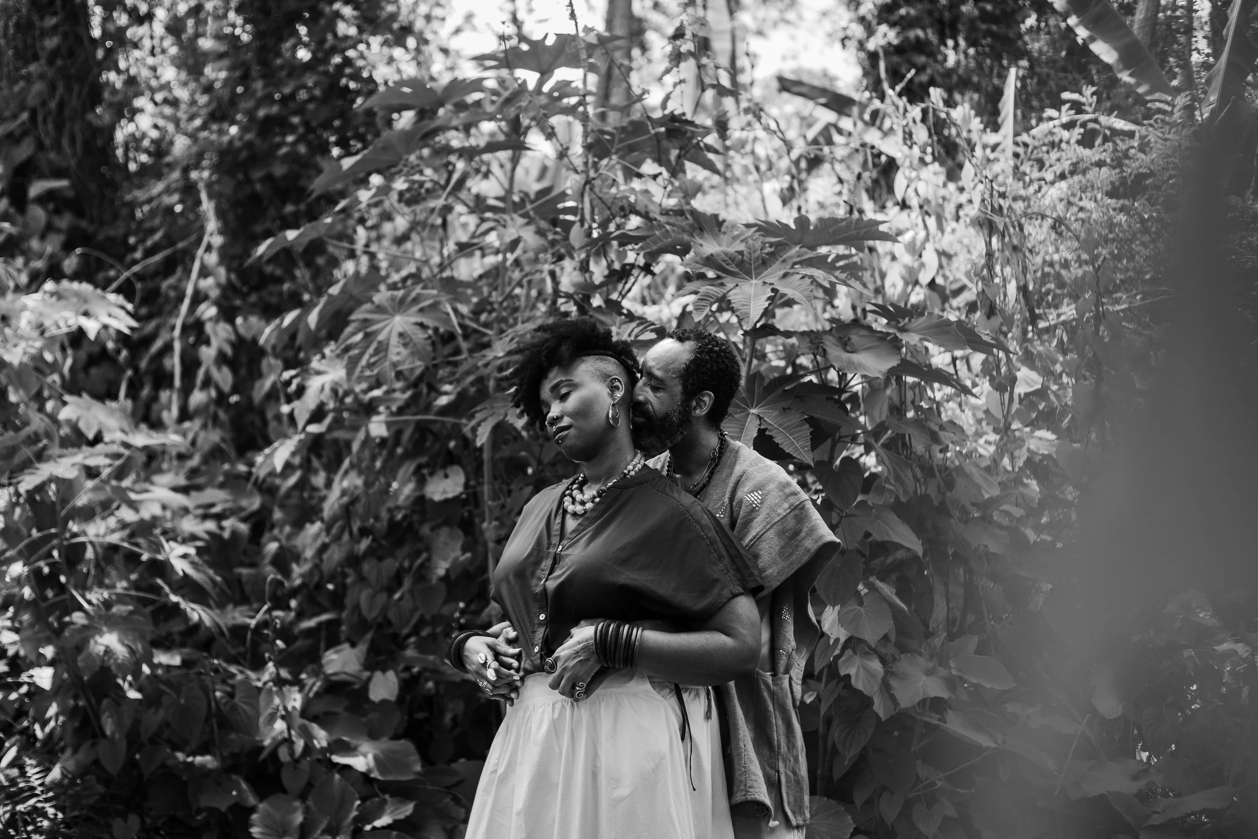 a black couple embracing from behind with their eyes closed surrounded by tropical plants.  The man is whispering in her ear.