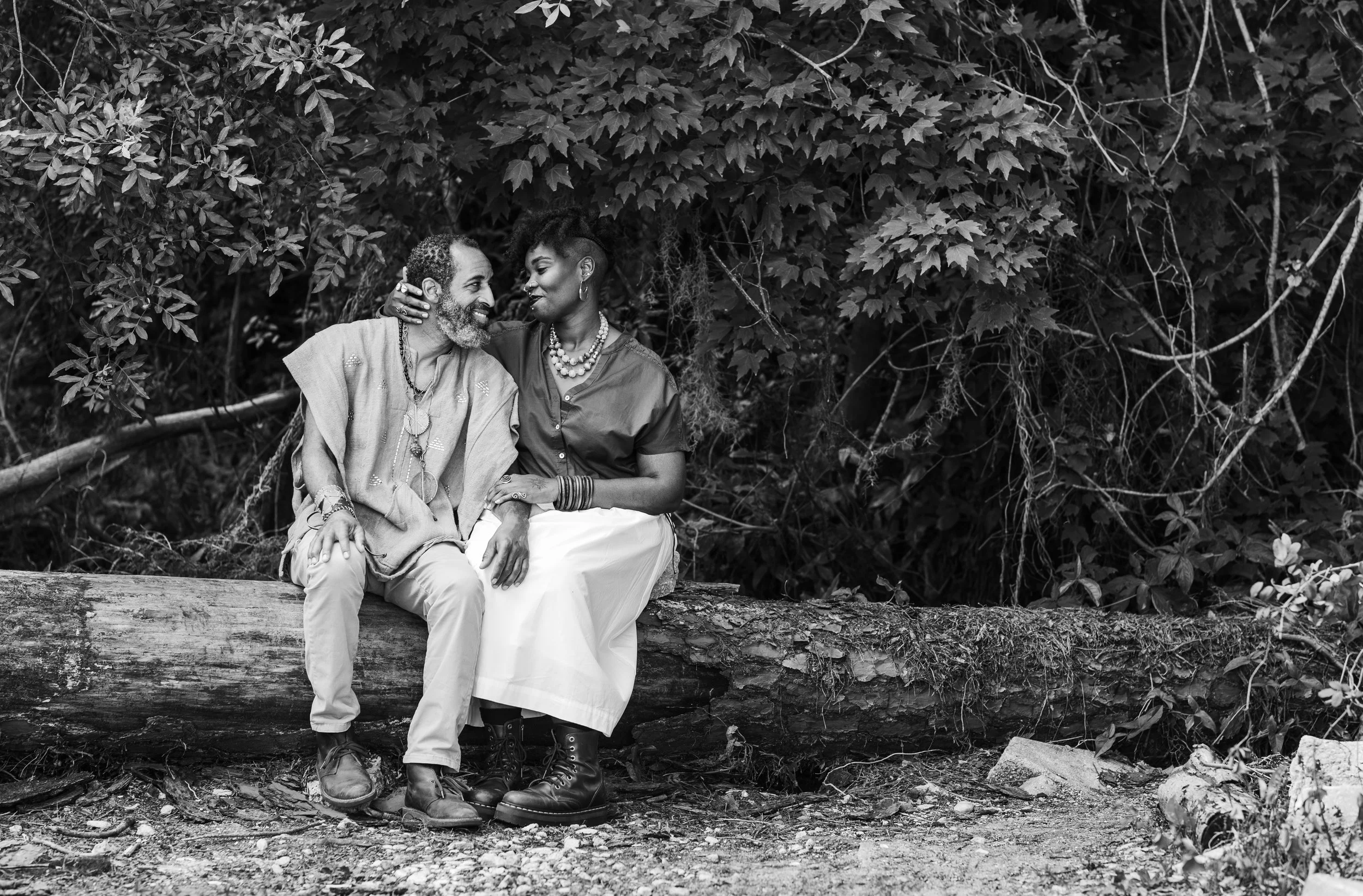 An engaged black couple sitting on a fallen tree trunk holding hands and looking into each others eyes