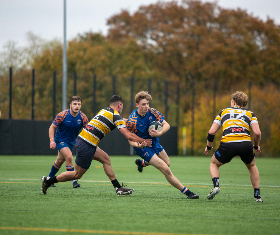 Rugby players in action on the field, with one player running and holding the ball while others attempt to tackle or block him, during a match with autumn trees in the background.