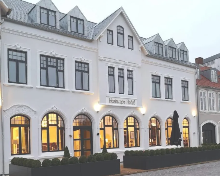 White multi-story building with large arched windows, sign reading 'Hobnobs Hotel,' and decorative exterior lighting, with a row of trimmed bushes in front.