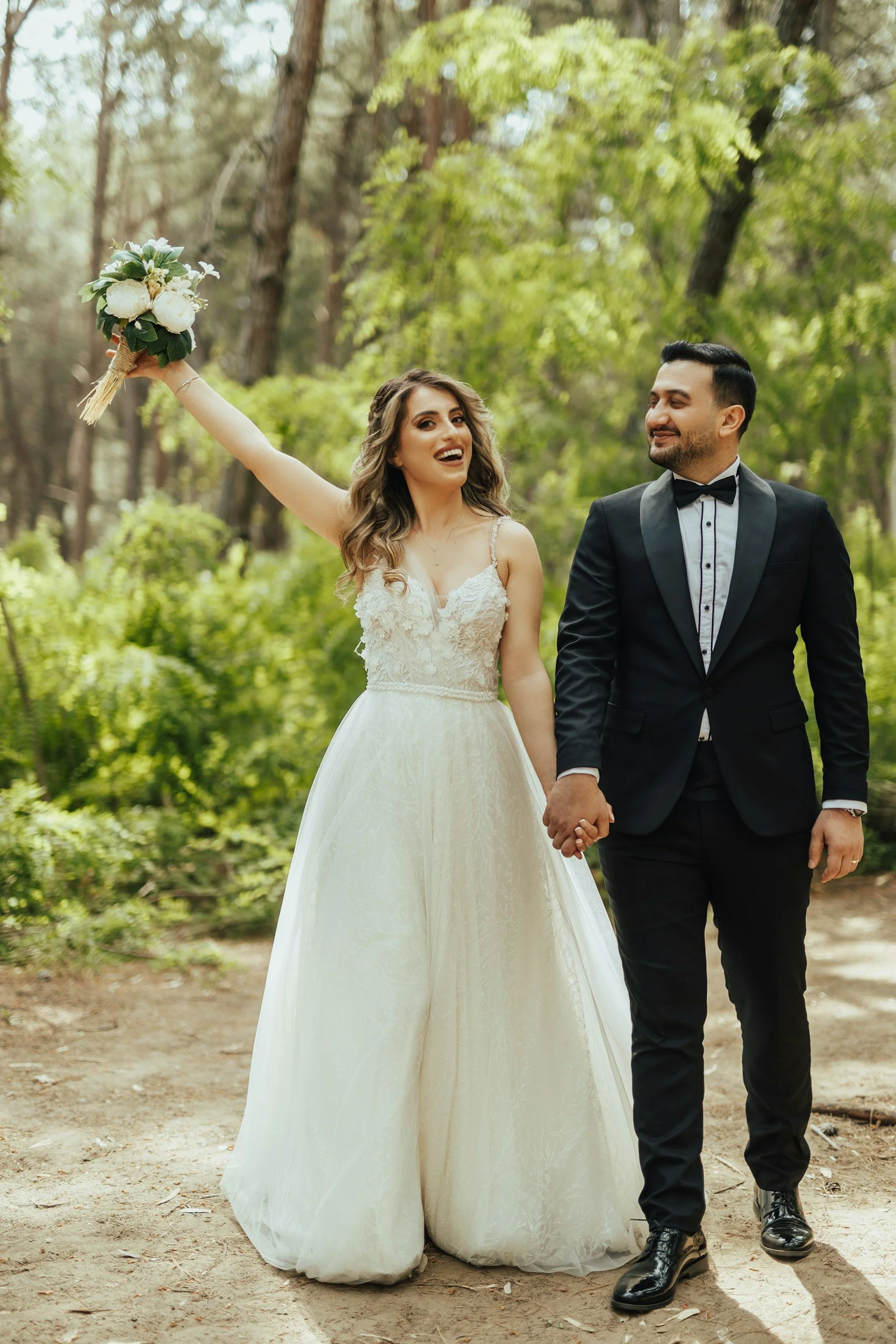 A newlywed couple walking hand in hand through a forest, the bride holding a bouquet up in the air and smiling, the groom looking at her lovingly, surrounded by green trees and sunlight filtering through the leaves.