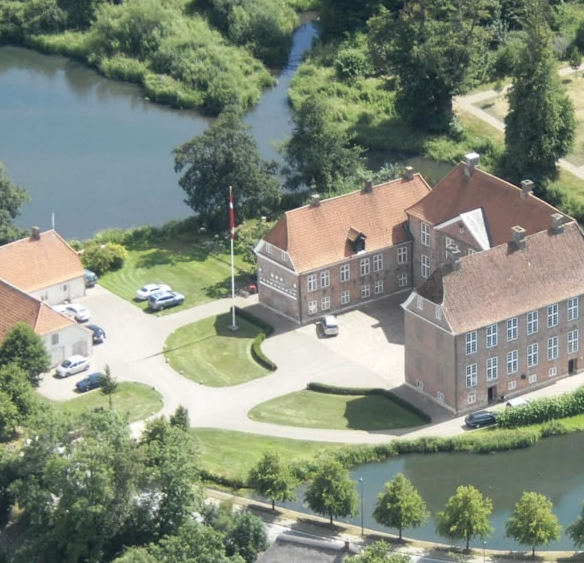 An aerial view of a large brick building with a red roof, surrounded by greenery and a pond, with a flagpole and parking lot nearby.