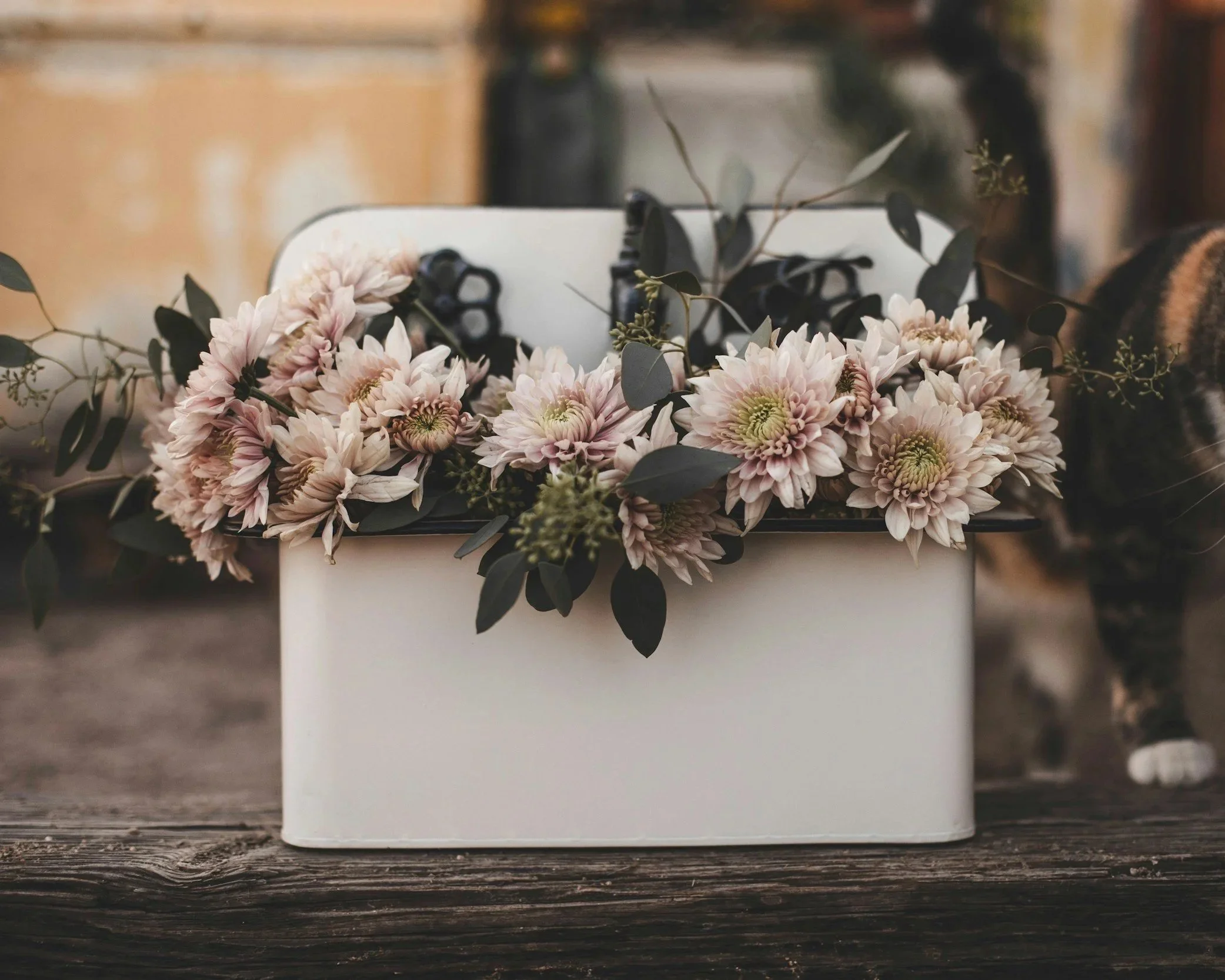 A white rectangular flower box filled with pink and white chrysanthemums and eucalyptus leaves, placed on a rustic wooden surface.