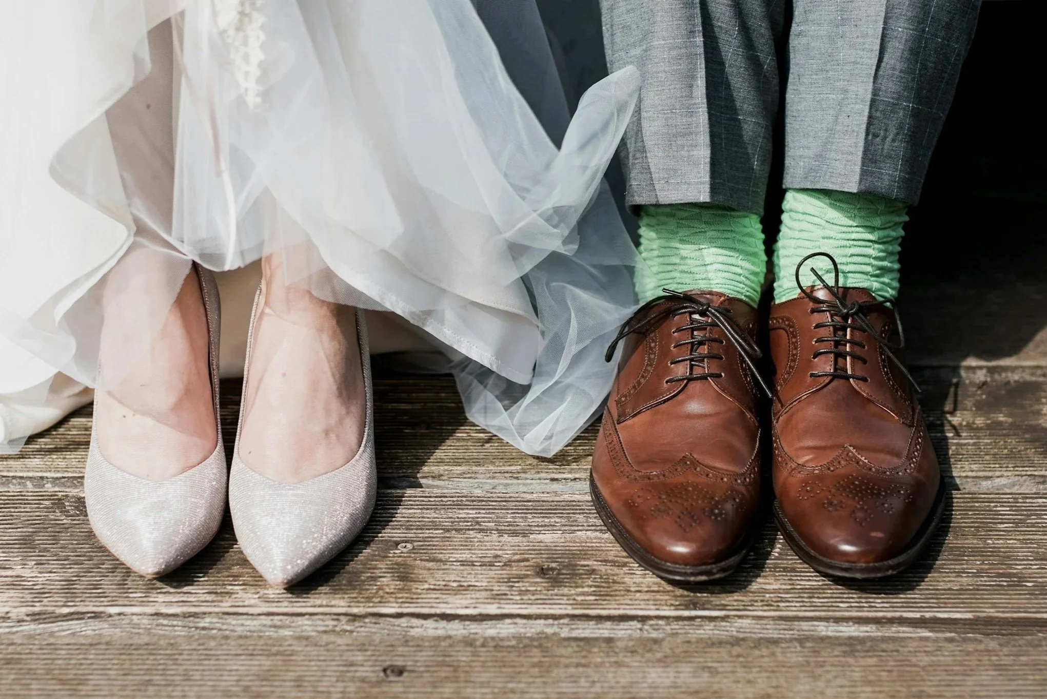 Close-up of a bride's white shoes next to a groom's brown shoes and bright green socks on a wooden floor.