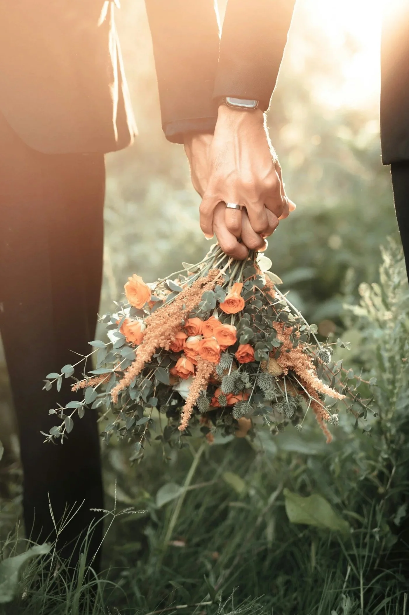 Close-up of two people holding a bouquet of orange flowers and greenery outdoors, with sunlight filtering through.