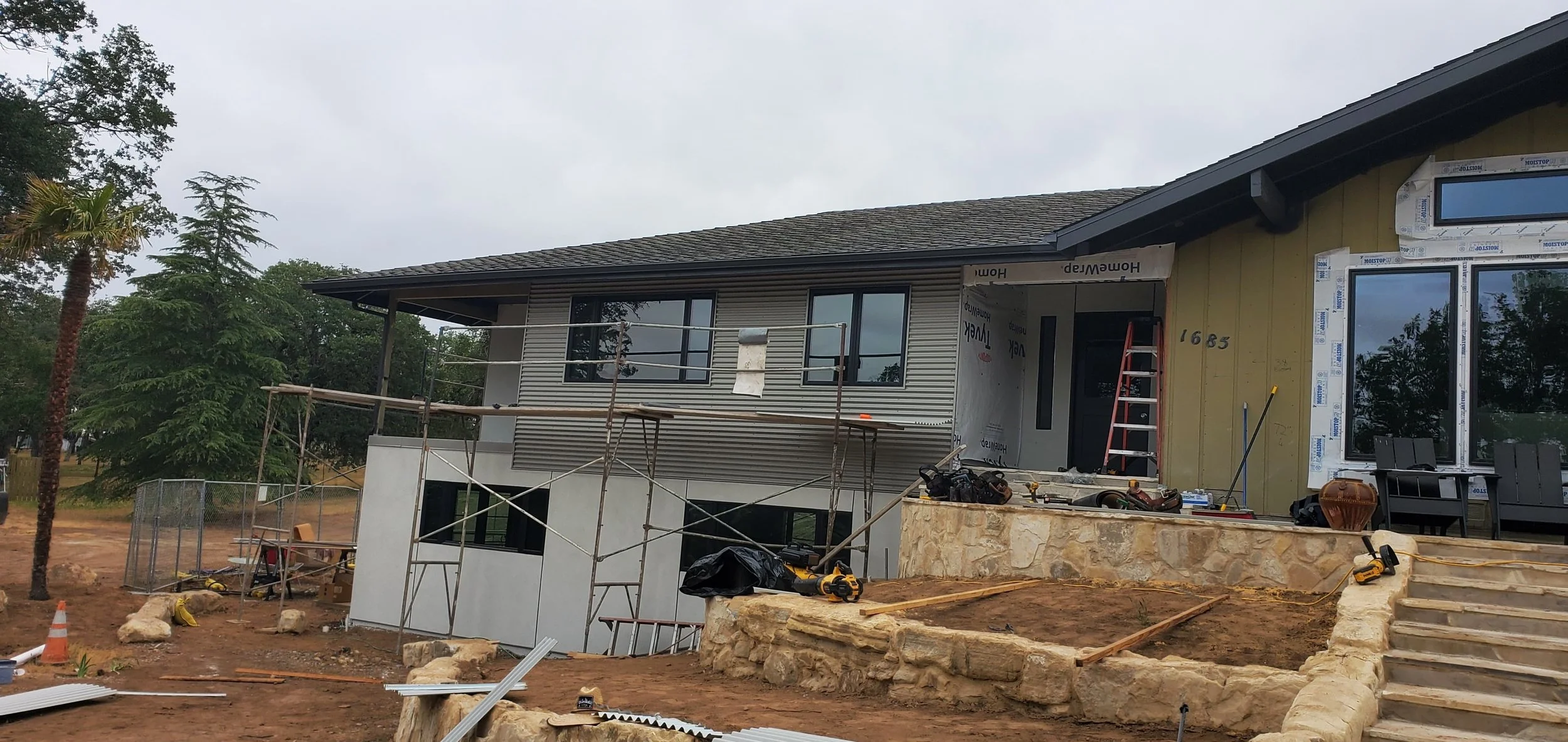A modern house sits on a grassy hill with mountain ranges in the background, some peaks snow-capped, under a cloudy sky.