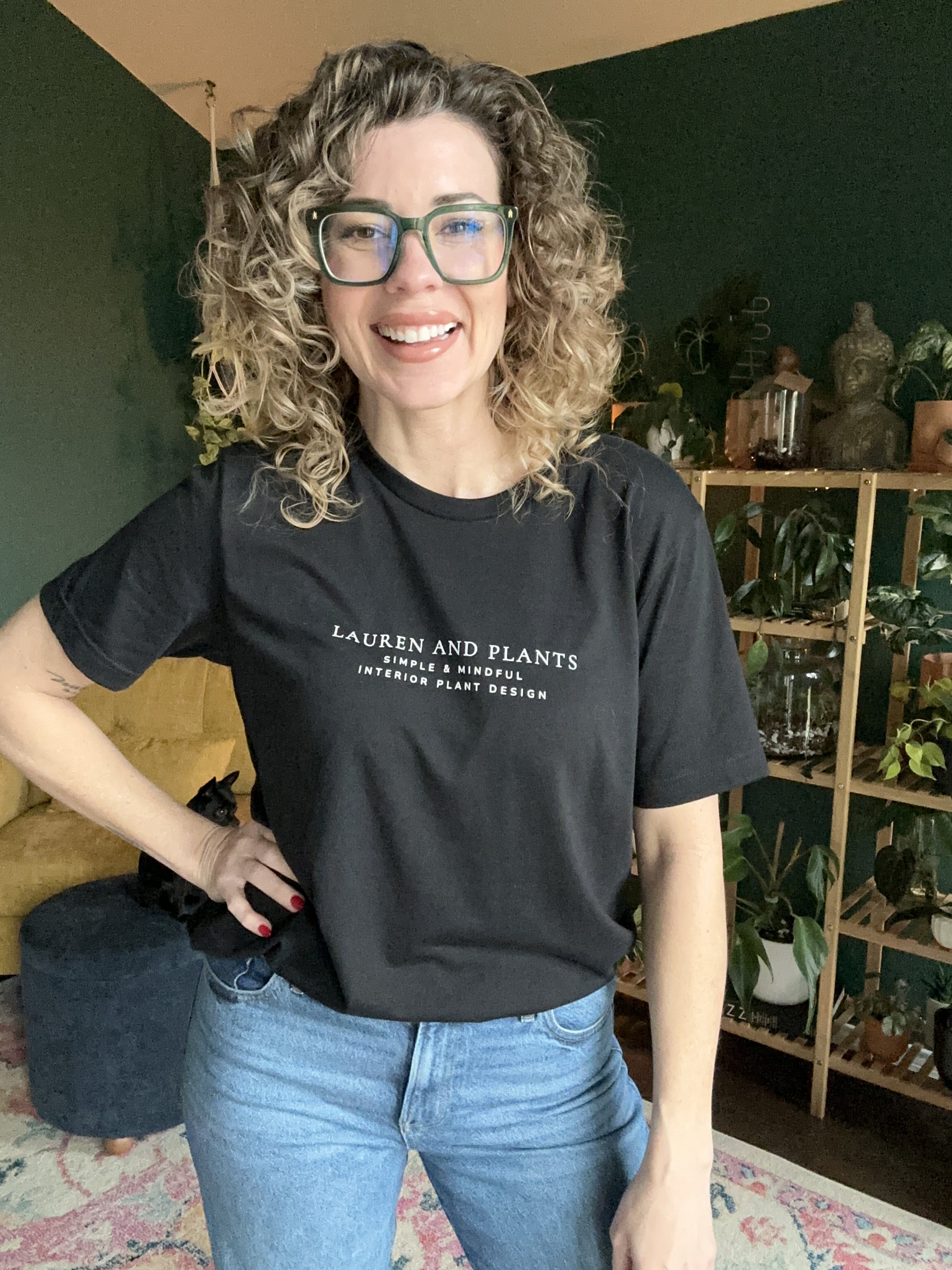 Lauren White, owner of Lauren and Plants, smiles in her plant room while wearing a Lauren and Plants shirt. The photo highlights her cozy, simple, and mindful plant design brand based in Fort Worth, Texas.
