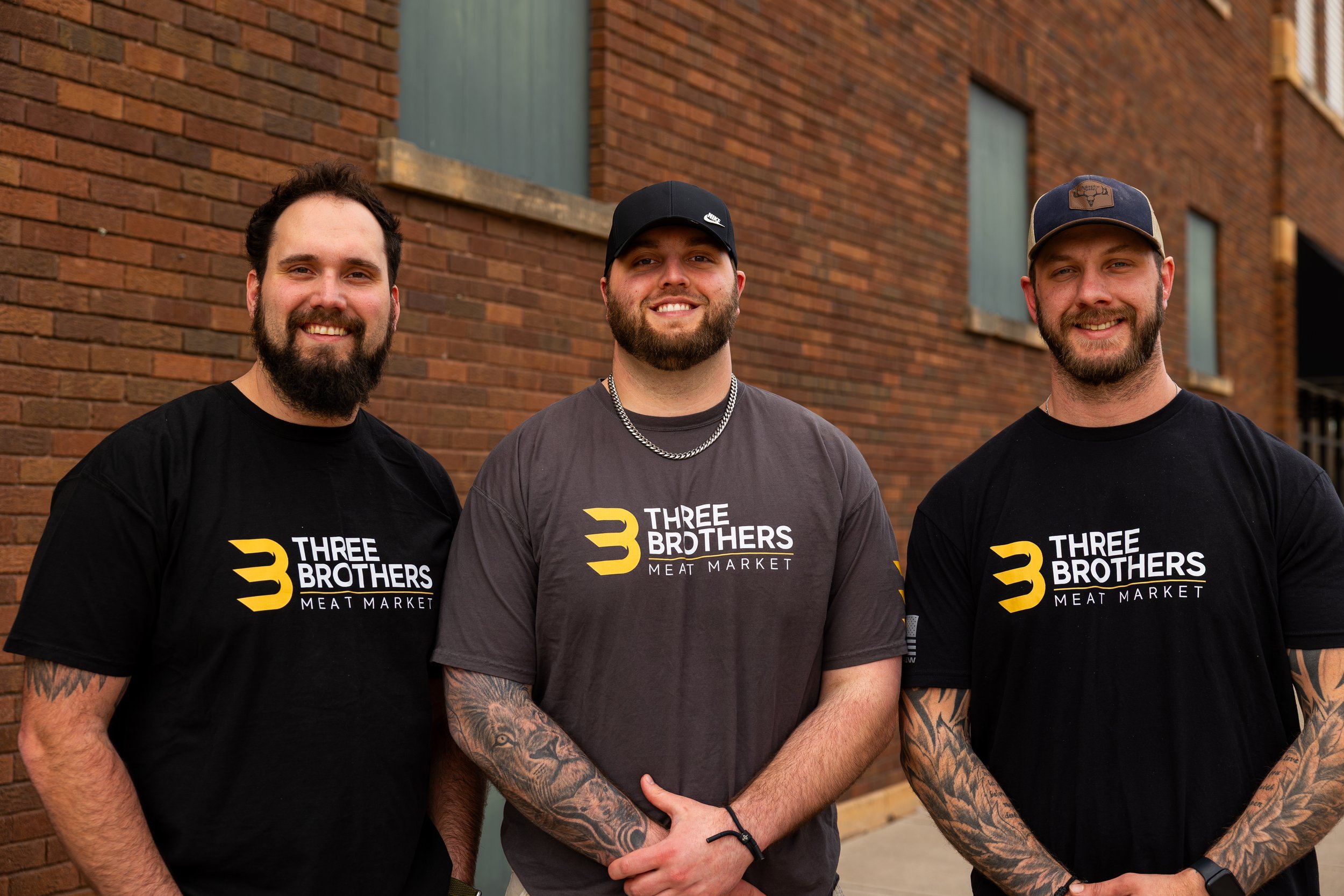 Three bearded men standing outdoors in front of a brick building, smiling and wearing T-shirts with the logo 'Three Brothers Meat Market'.