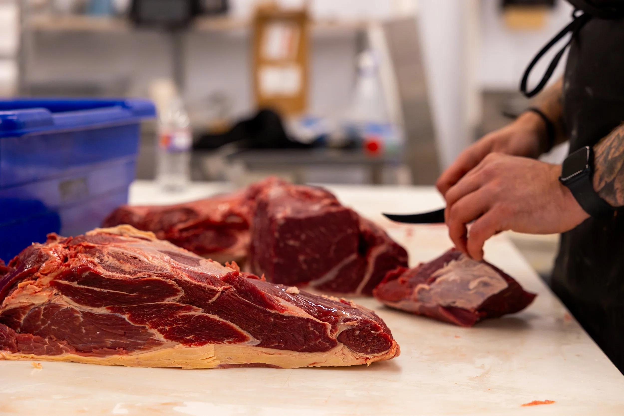 Person using a knife to cut large raw beef steaks on a white cutting board in a kitchen or butcher shop.