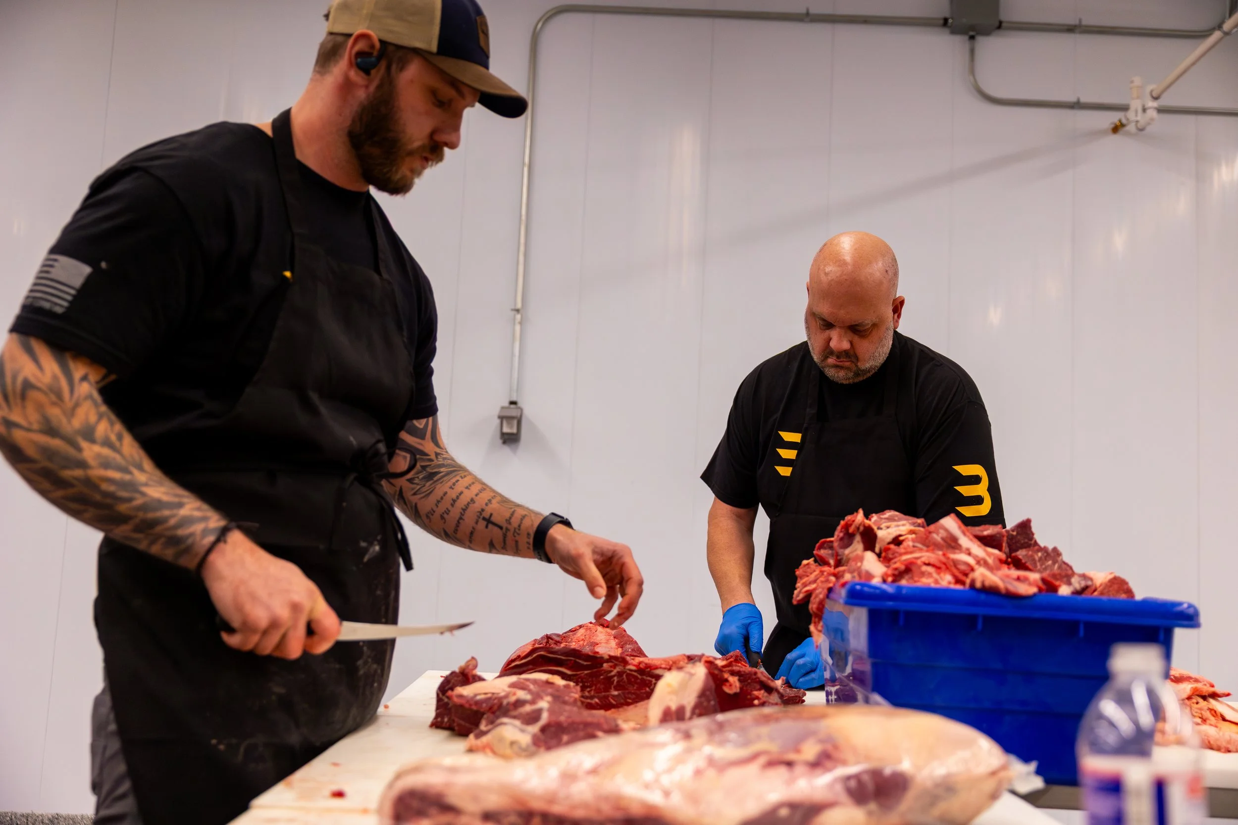 Two men butchering and preparing raw meat in a meat processing facility or butcher shop, with large cuts of meat on the table and a blue container filled with meat.