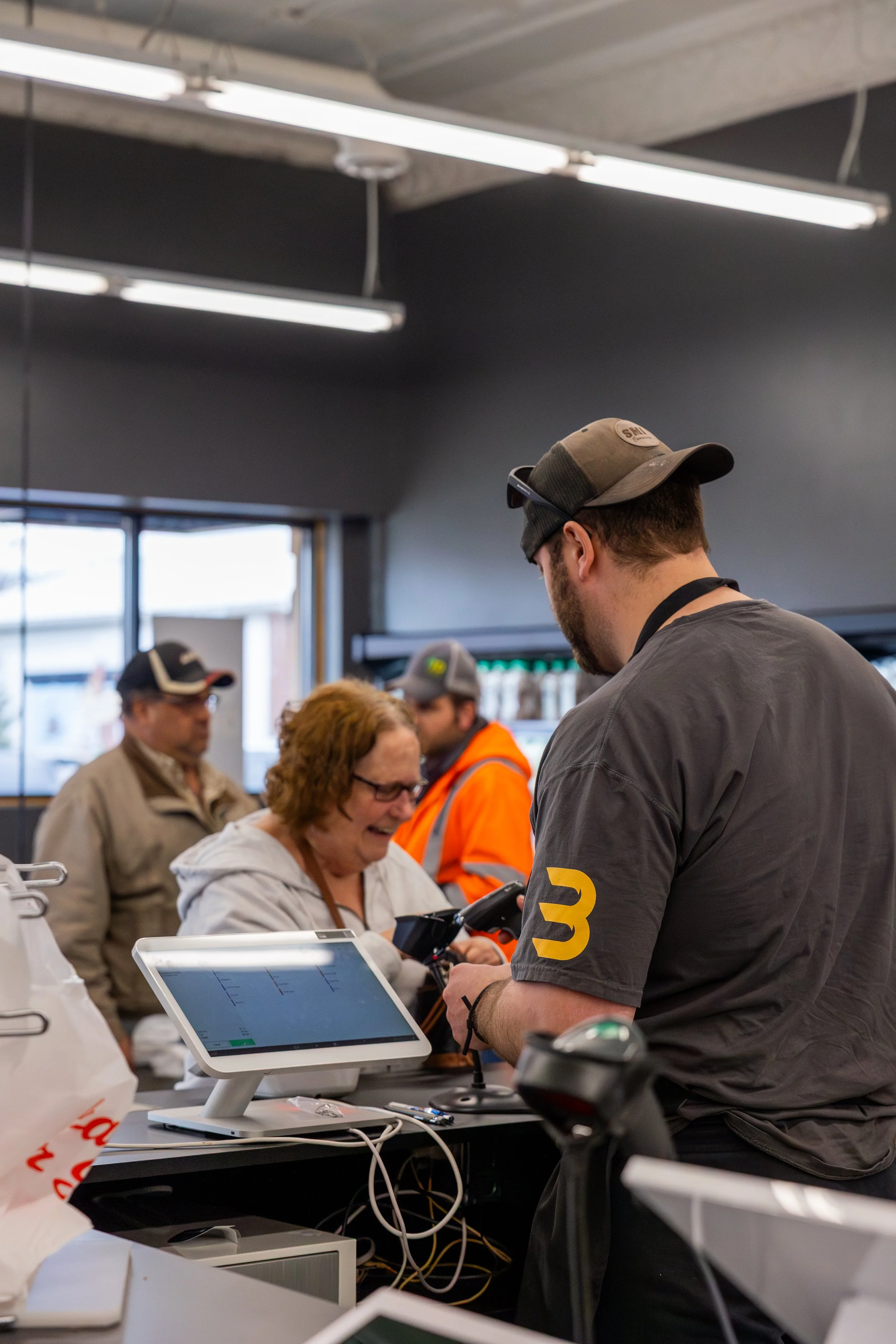 Employees at a checkout counter assist a smiling customer, with store shelves visible in the background.
