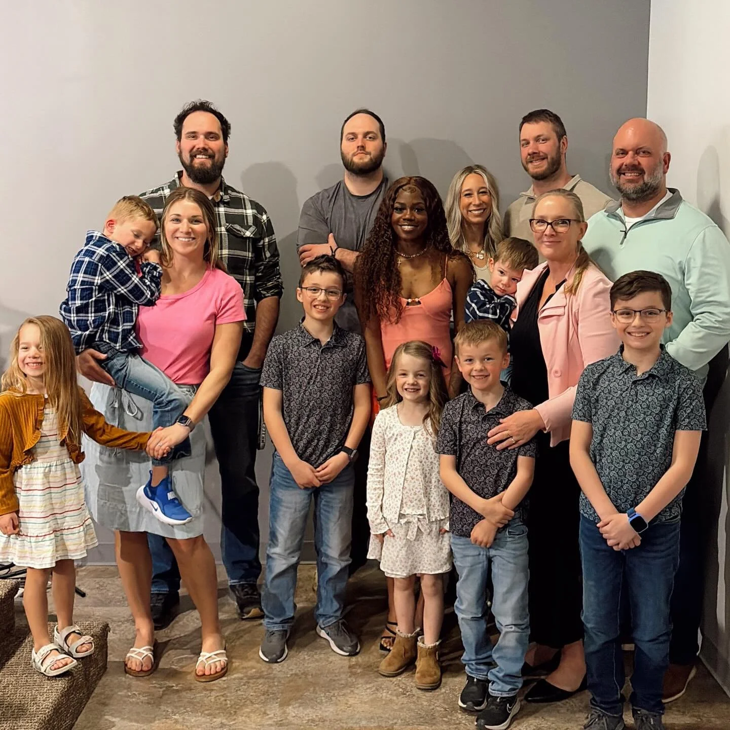 A large family group photo with adults and children posing indoors against a plain wall.