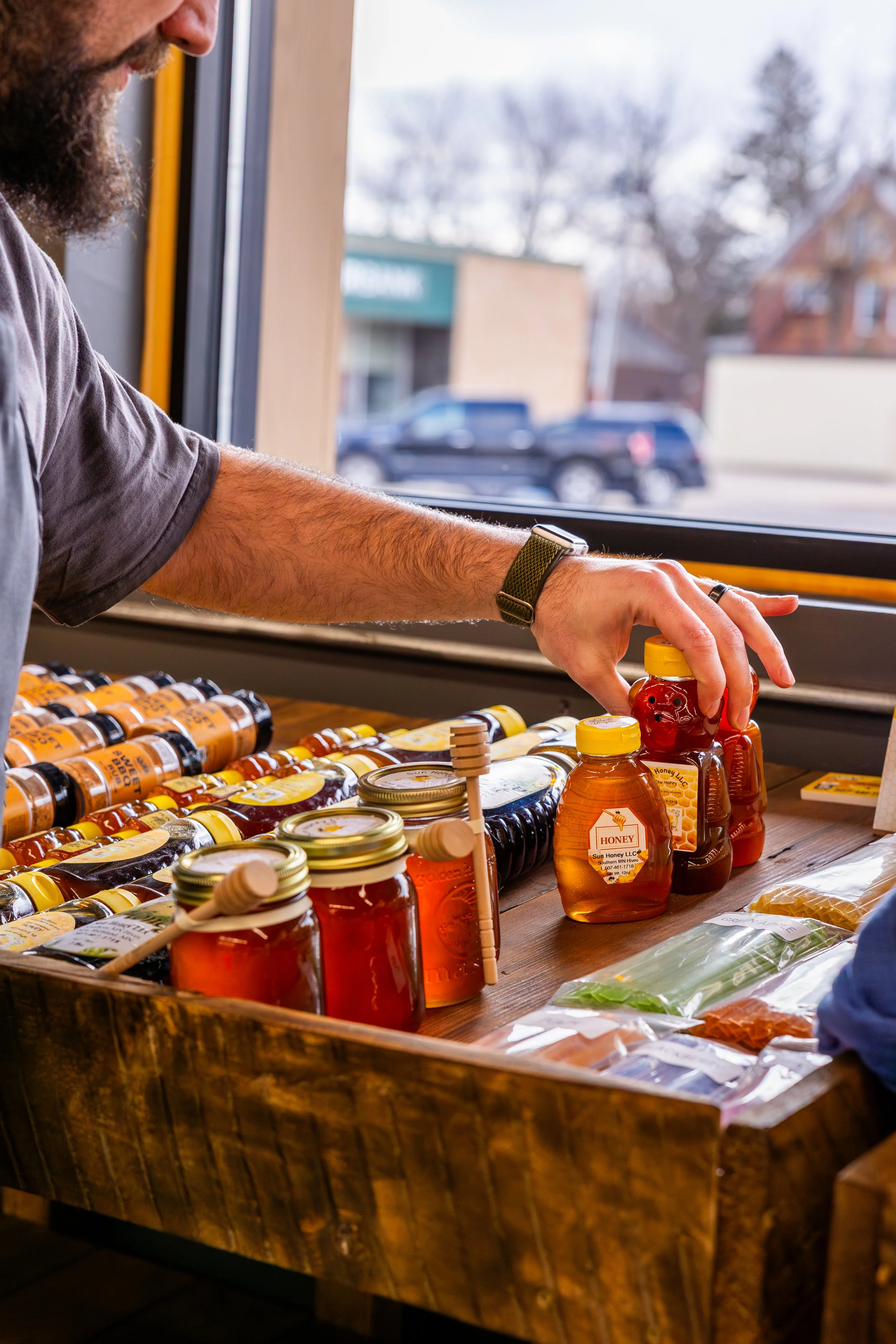 Person reaching for honey bottles at a market stand with jars and bottles of honey inside.