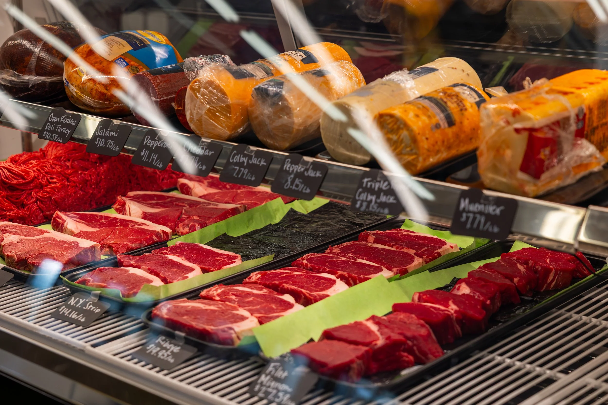 A display case of various cuts of raw beef in a grocery store, with cheese and packaged meats on the top shelf.