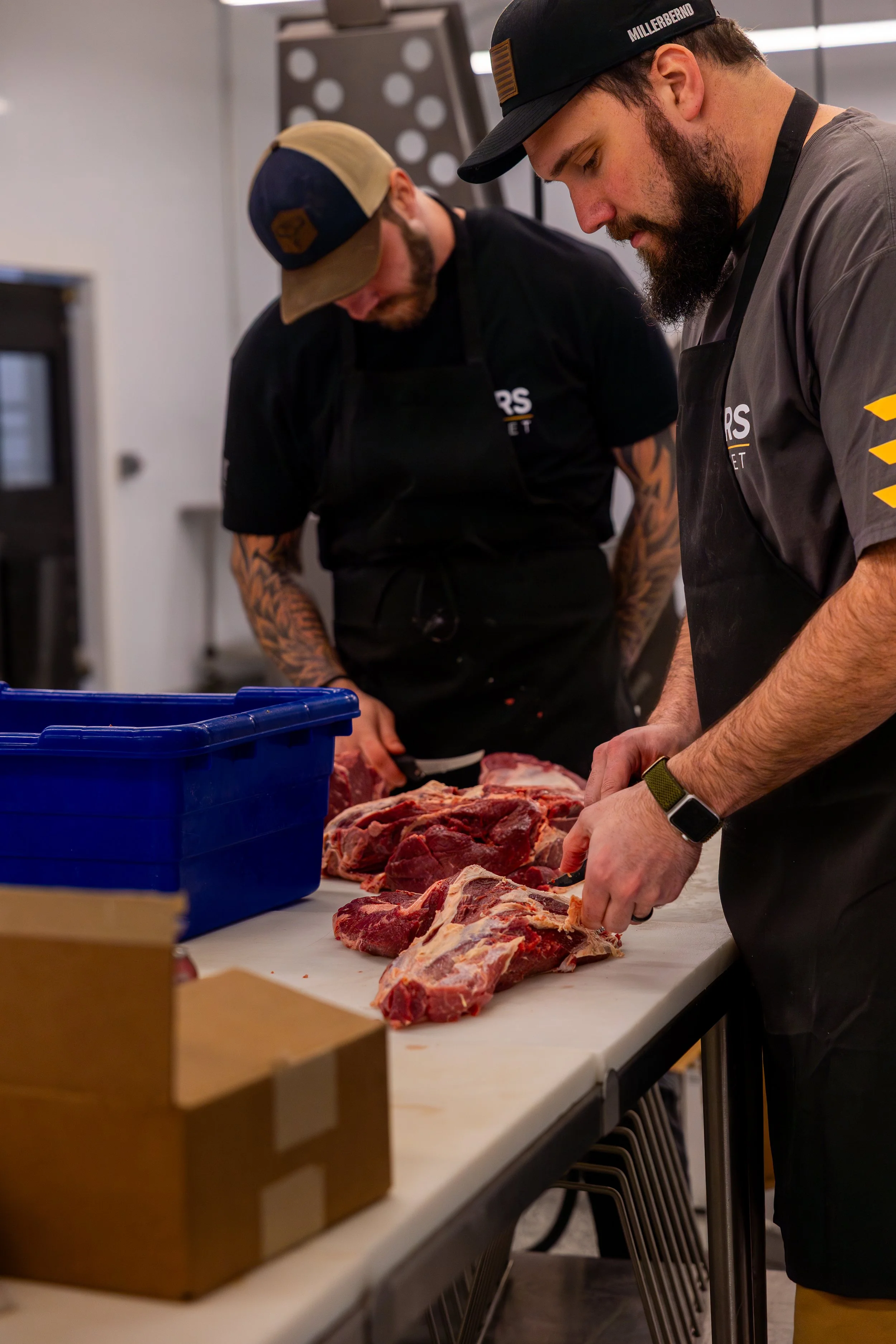 Two men in black and gray aprons and hats are cutting raw meat on a white table in a professional kitchen or butchery setting.