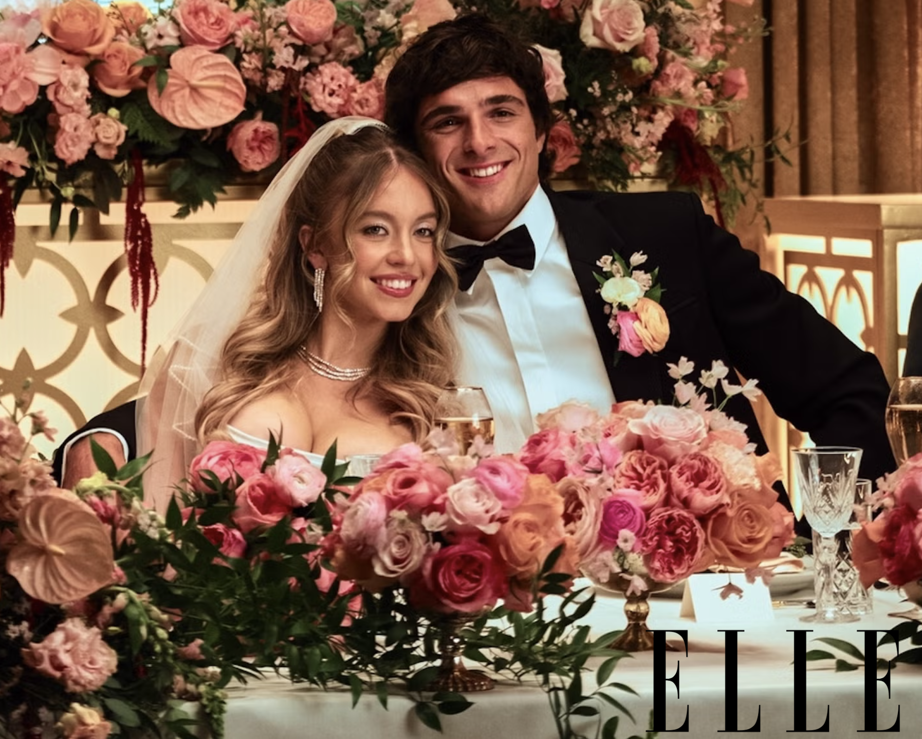 A bride and groom at their wedding reception sitting behind a large floral centerpiece. The bride has long wavy blonde hair, is wearing a white gown with off-the-shoulder sleeves, pearl jewelry, and a veil. The groom with dark hair is dressed in a bl