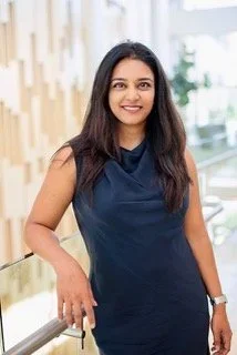 A woman with long dark hair smiling, wearing a navy sleeveless top, standing indoors near a railing with a modern interior background.