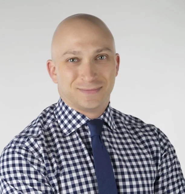 A young man with a shaved head, wearing a blue and white checkered shirt and a dark blue tie, smiling against a plain light gray background.