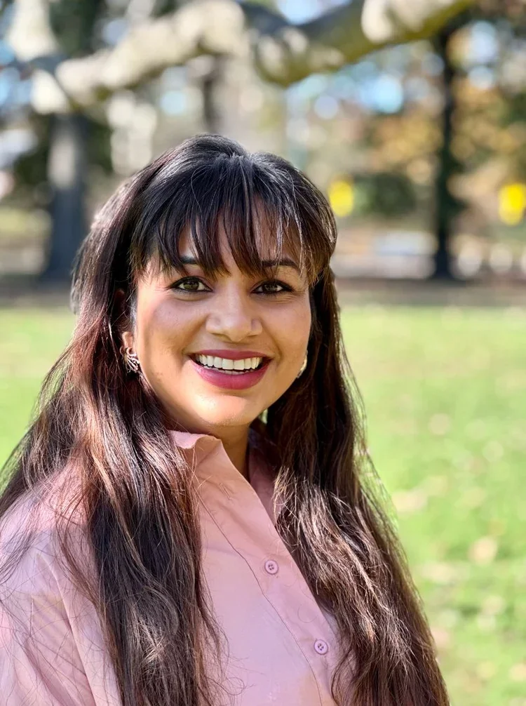A woman with long brown hair and bangs smiling outdoors on a sunny day.