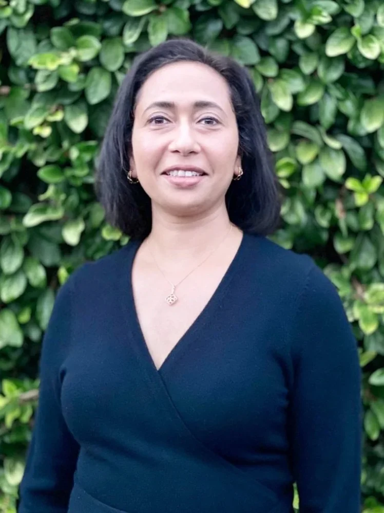 A woman with shoulder-length black hair, wearing a black top and jewelry, standing in front of green foliage.