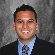 Headshot of a man with dark hair, wearing a black blazer, light blue shirt, and a patterned tie, smiling against a gray textured background.
