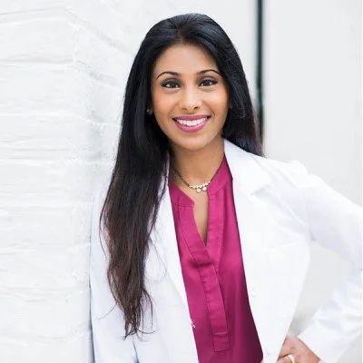 A young woman with long dark hair, smiling, wearing a white coat over a pink top, standing next to a white wall.