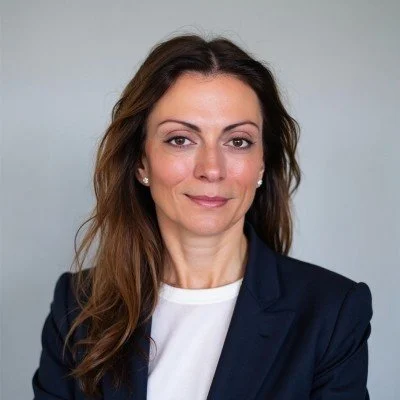 Professional woman with long brown hair, wearing a navy blazer and white top, posing against a neutral background.