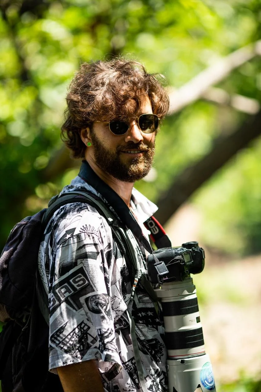 A man with curly brown hair, sunglasses, and a beard stands outdoors in a green, leafy environment. He is wearing a patterned shirt and carrying a backpack with a camera hanging from his neck.